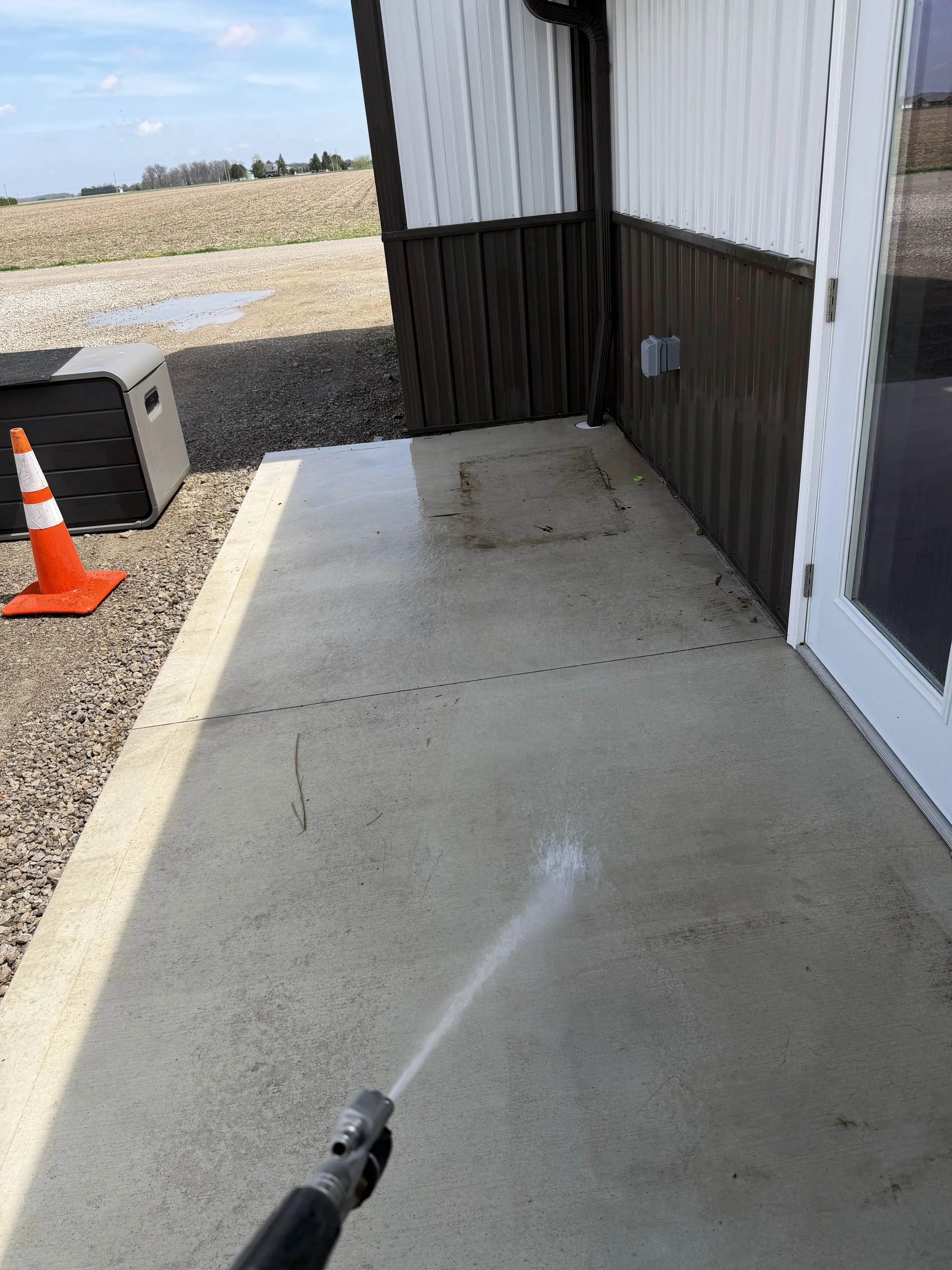 A person pressure washing a concrete patio outside a building with a metal siding, with an open landscape and blue sky in the background.