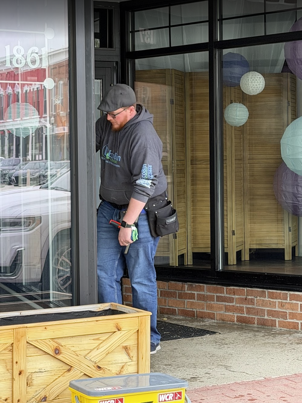 A man with glasses, a black cap, gray hoodie, and jeans is standing outside a storefront with large glass windows, looking down at something. He has a tool belt on his waist and is holding a green and black object in his left hand. There is a wooden planter box and a yellow box labeled "WCR" on the ground in front of him, with a background of parked cars and buildings.
