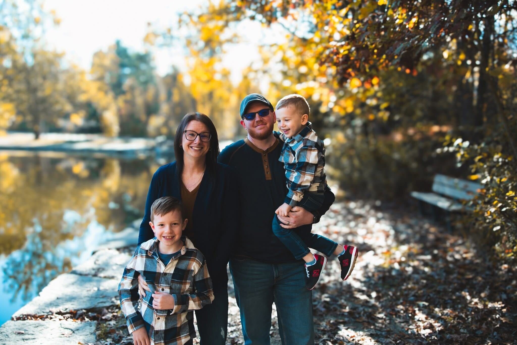 A family of four standing outdoors near a water body in autumn, smiling and enjoying the weather. They are dressed casually, with the adults wearing glasses and one man wearing sunglasses and a cap. The background features trees with fall foliage.