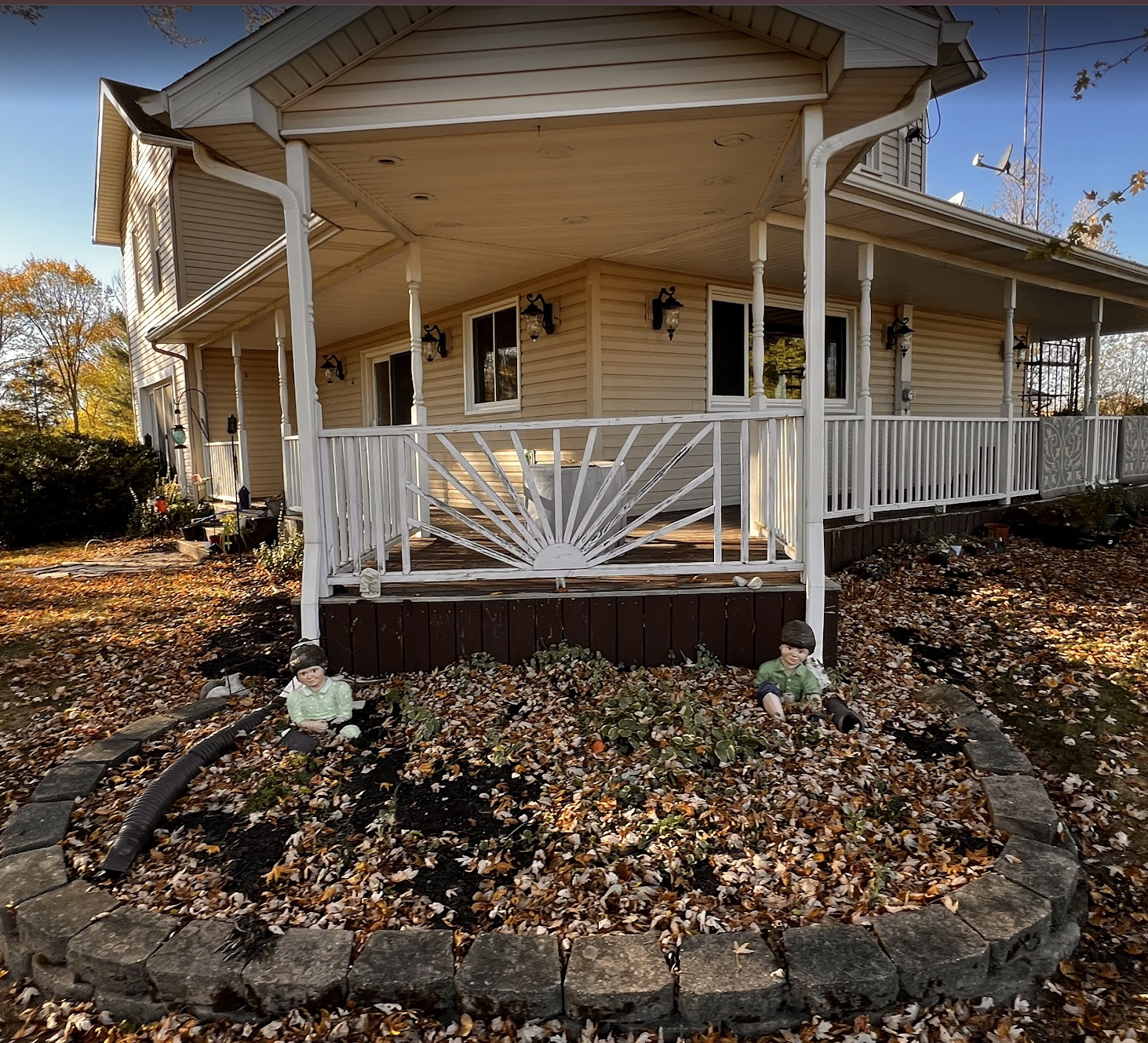 A house with a wraparound porch and white railing, surrounded by fallen autumn leaves. In the foreground, there's a flower bed with two small garden statues of children, one sitting and the other kneeling, planted among fallen leaves.