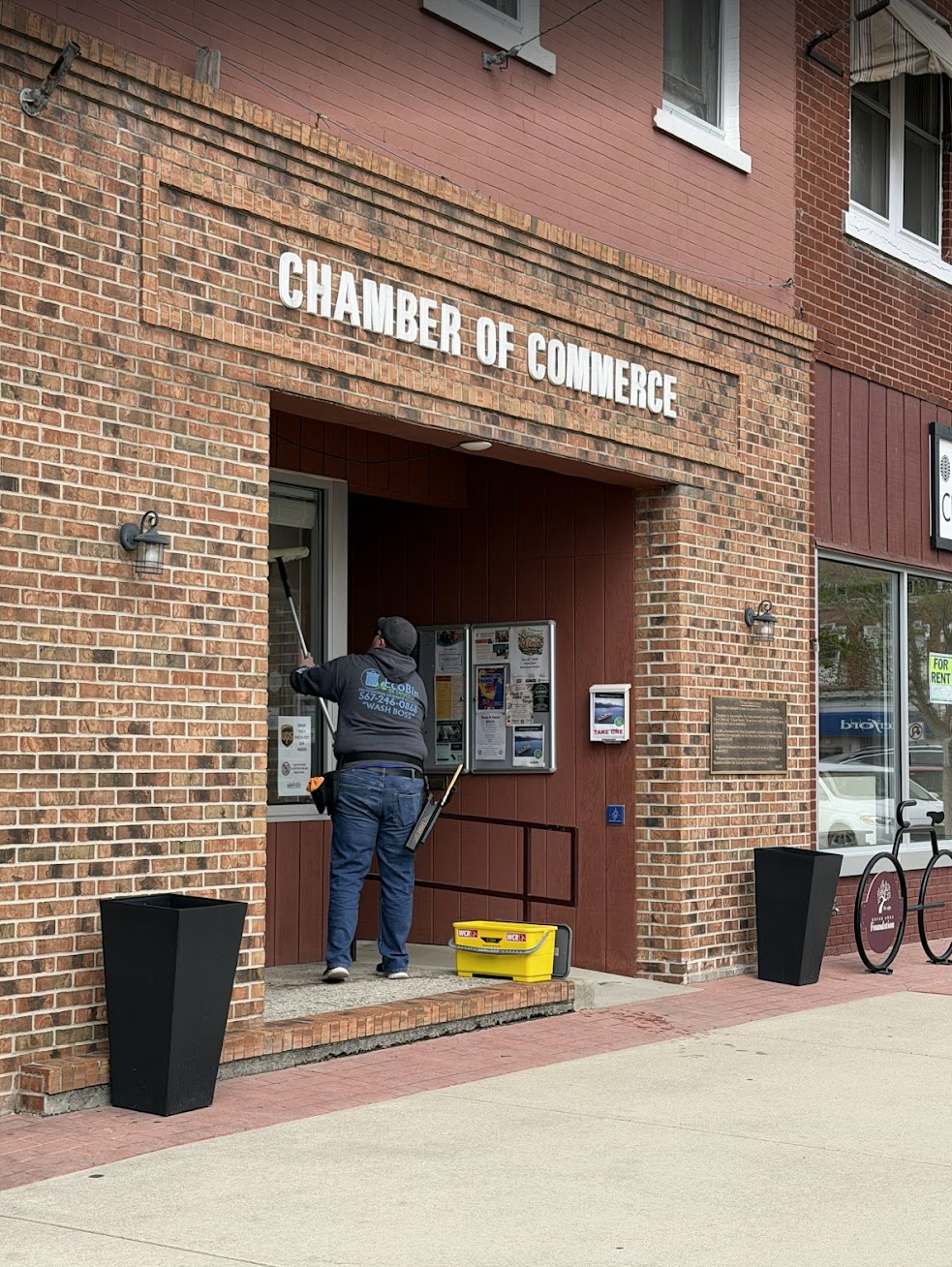 Person cleaning the windows at the Chamber of Commerce building with equipment on a sidewalk in a small commercial area.
