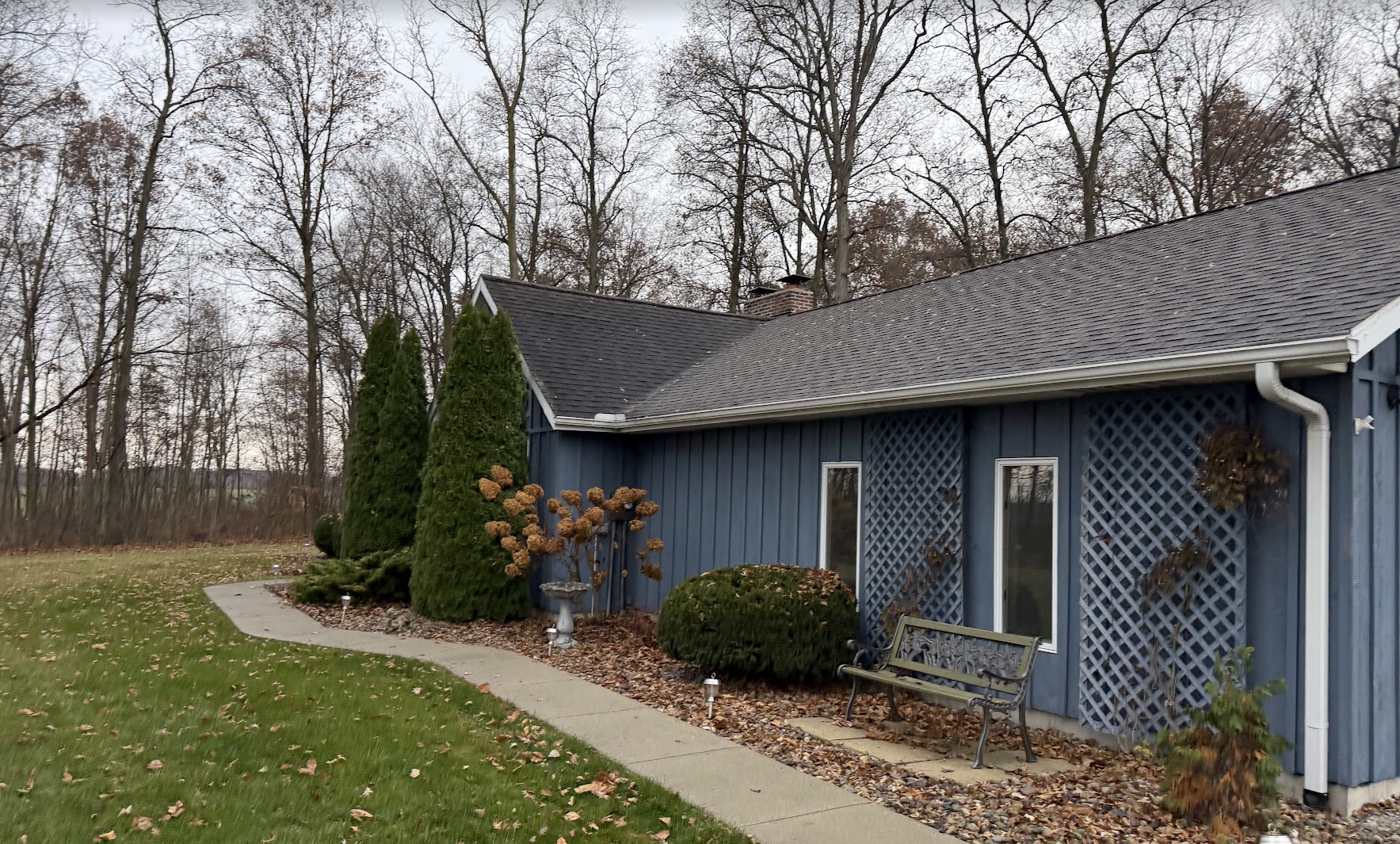 A house with dark blue siding and a sloped roof surrounded by leafless trees in a backyard garden with trimmed bushes, a concrete pathway, and a bench.