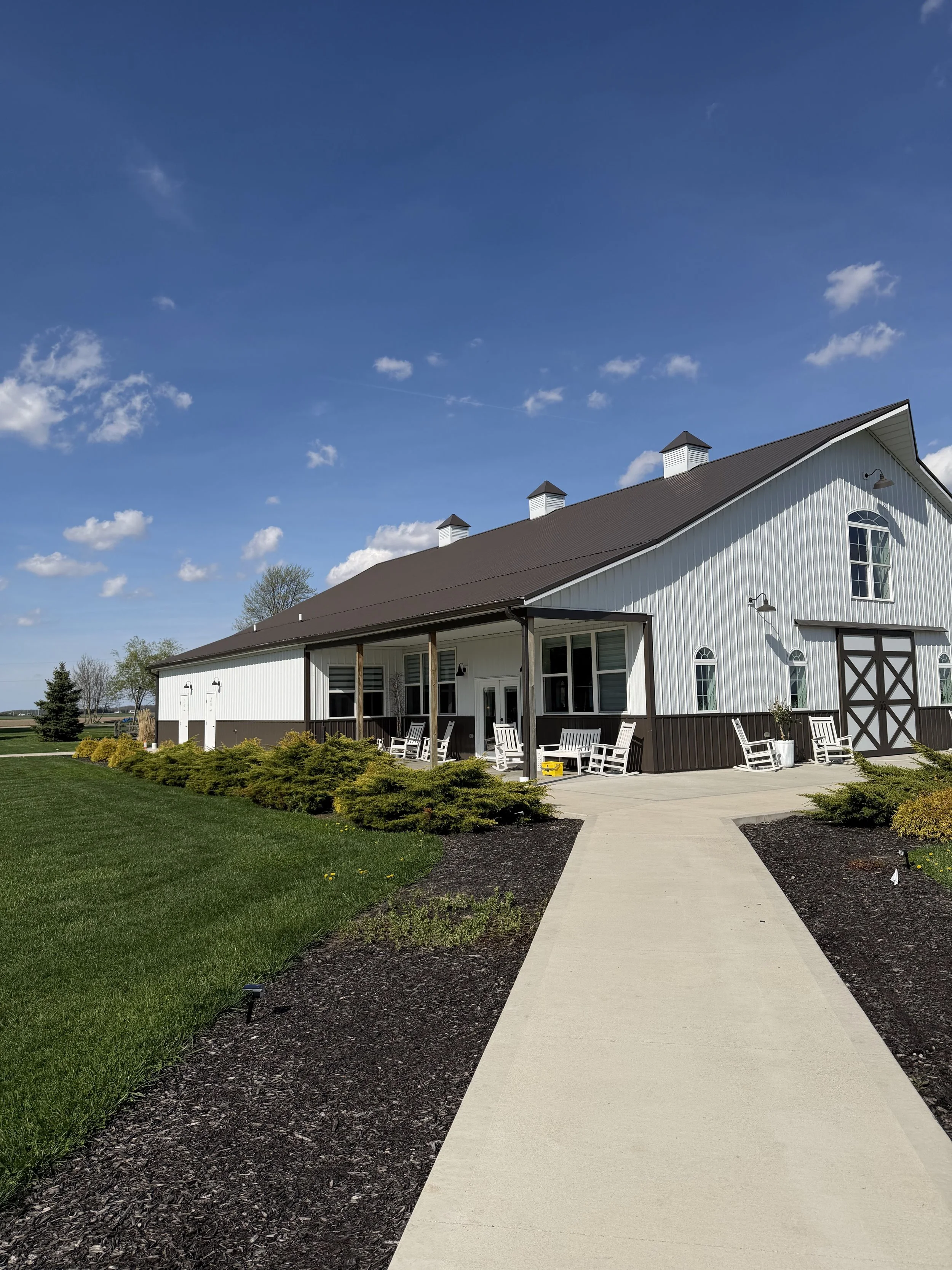 A white barn with black accents and a black metal roof, surrounded by a well-maintained lawn and a concrete pathway, under a bright blue sky with scattered clouds.