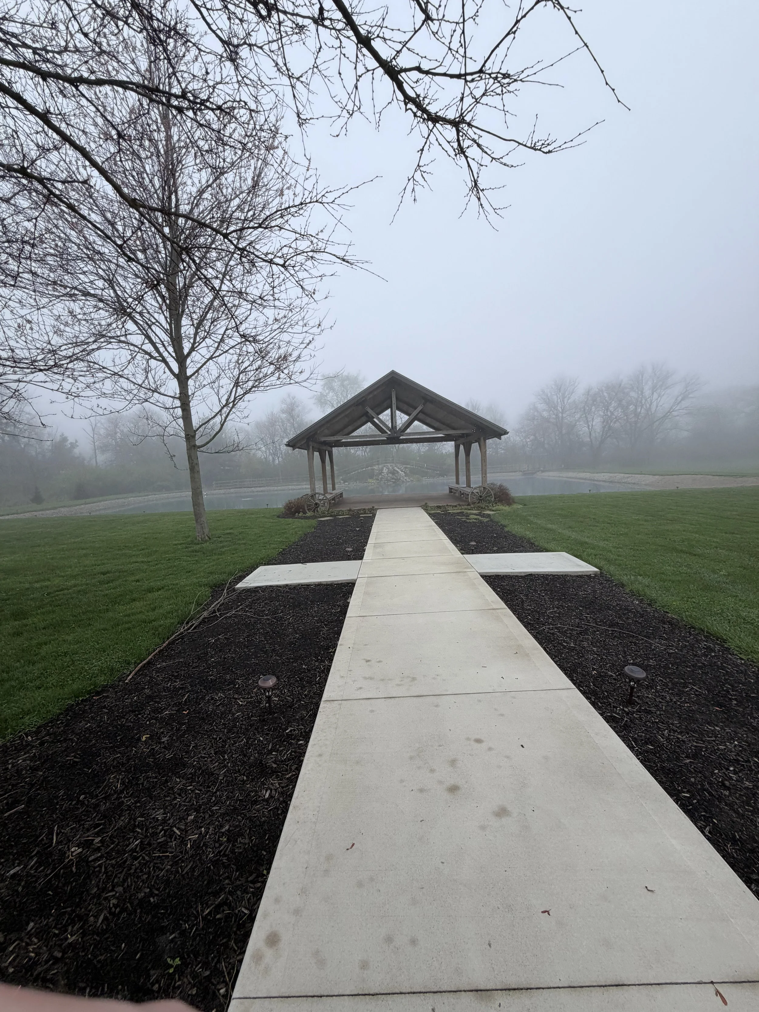 A foggy outdoor scene with a small wooden pavilion over a pond, trees around, and a paved walkway leading to the pavilion, with grass on either side.