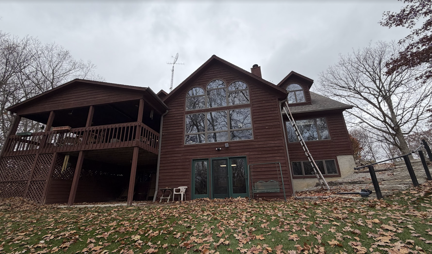 Back view of a two-story wooden house with a large arched window, smaller windows, a screened porch, and a ladder leaning against the side during fall with bare trees and fallen leaves.