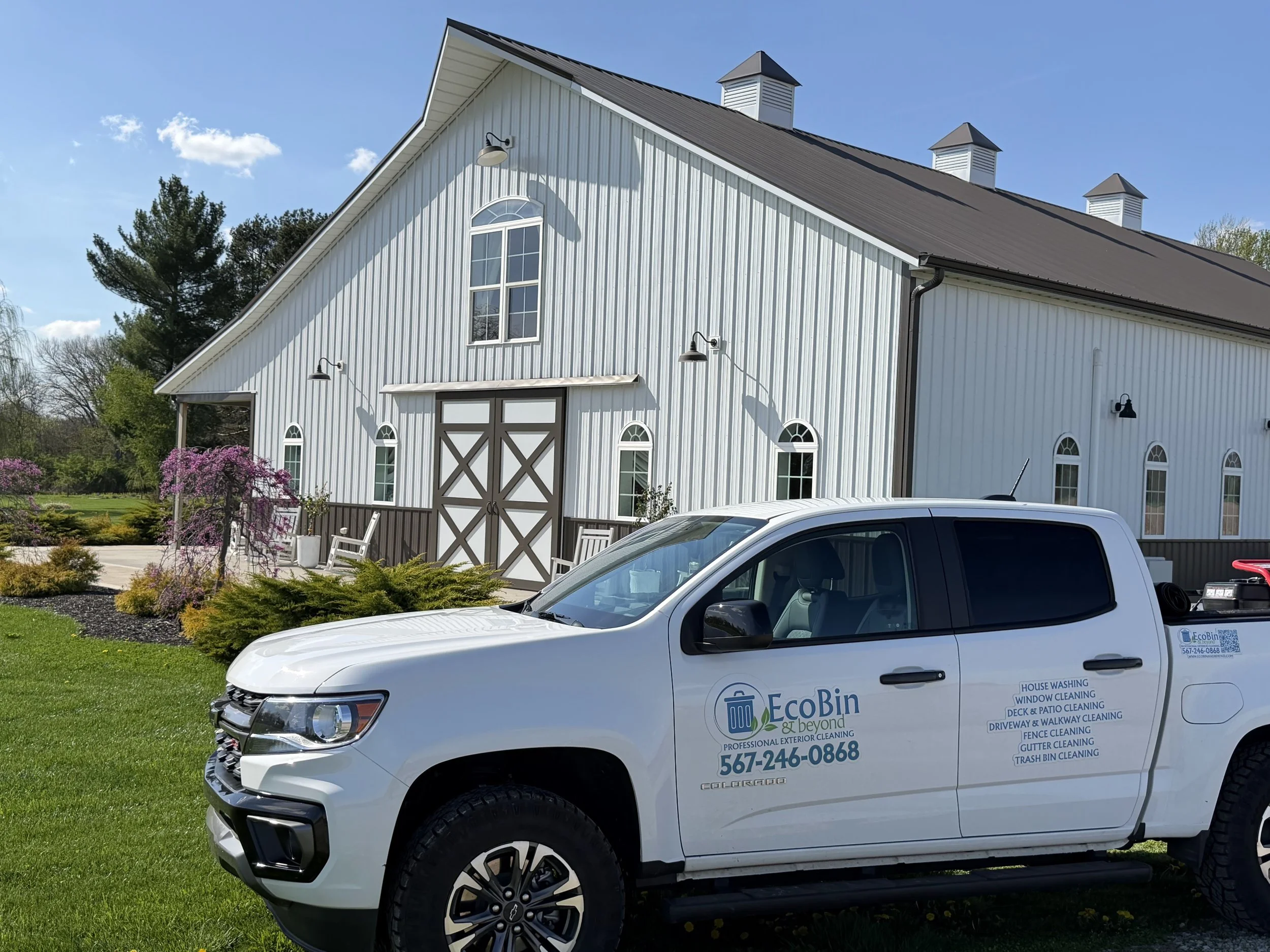 A white EcoBin vehicle parked on a well-manicured lawn in front of a large white barn with black roof accents, surrounded by trees and flowering bushes, on a sunny day.