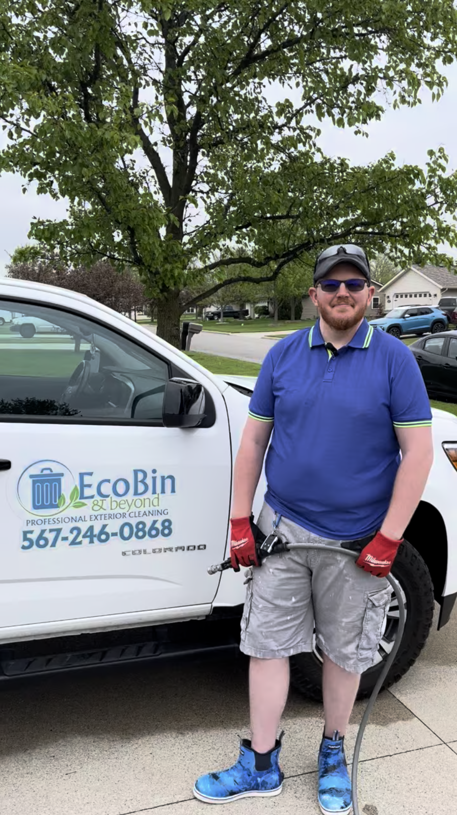 A man standing outdoors next to a white EcoBin service vehicle, holding a tool, wearing sunglasses, a cap, a blue polo shirt, beige cargo shorts, red gloves, and blue shoes, with green trees and a suburban neighborhood in the background.
