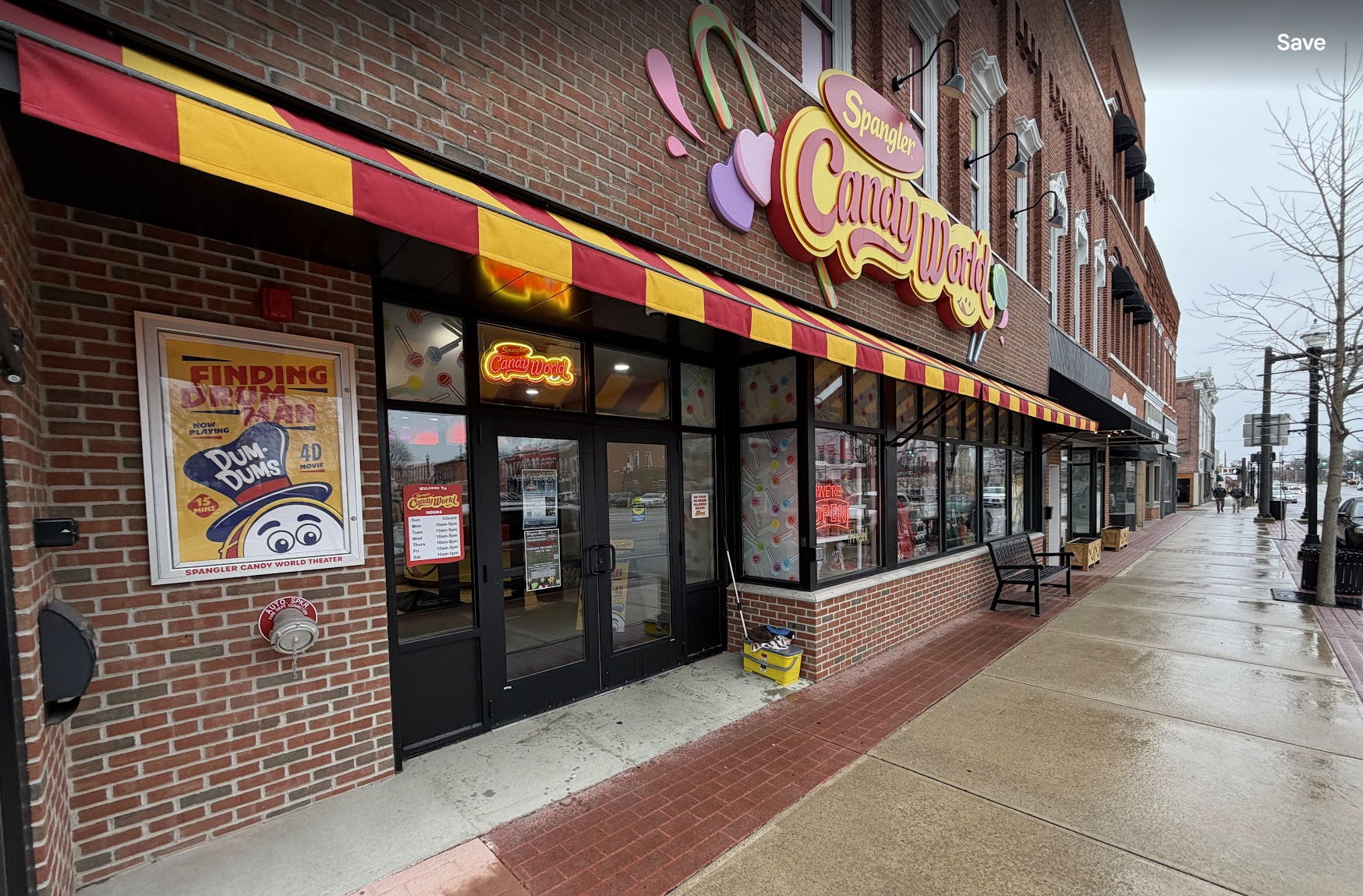 Exterior of a candy store called Spangler Candy World on a rainy day, with a brick building, colorful signage, a red and yellow striped awning, and a sidewalk with benches.