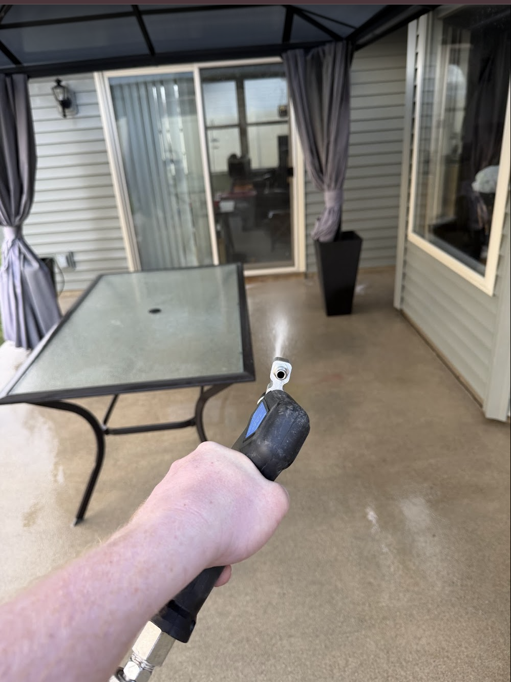 Person using a laser measuring tool on a covered outdoor patio with a glass table, sliding glass door, window, and potted plant.