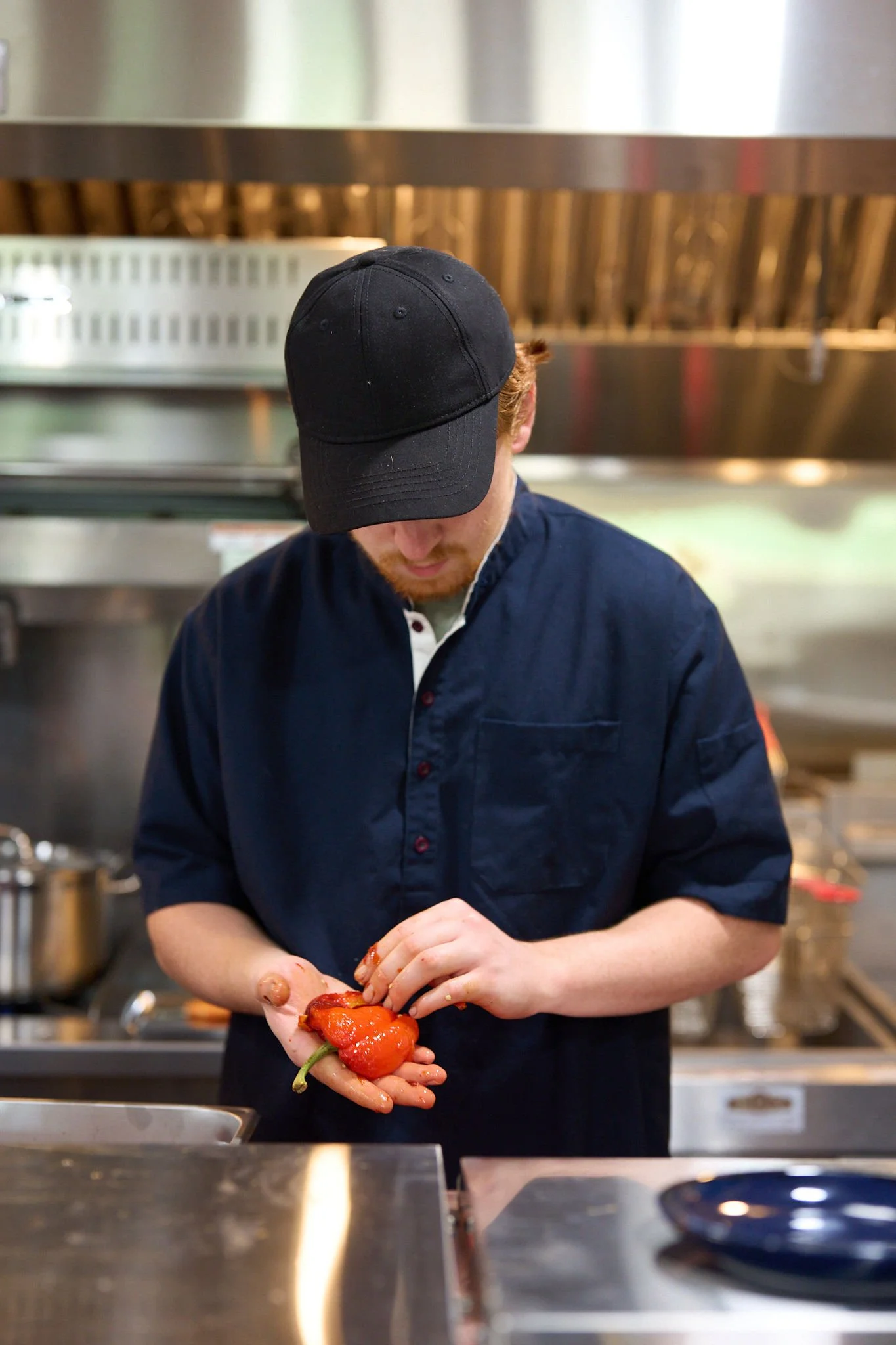 Person wearing a black cap and navy shirt preparing red peppers in a kitchen.