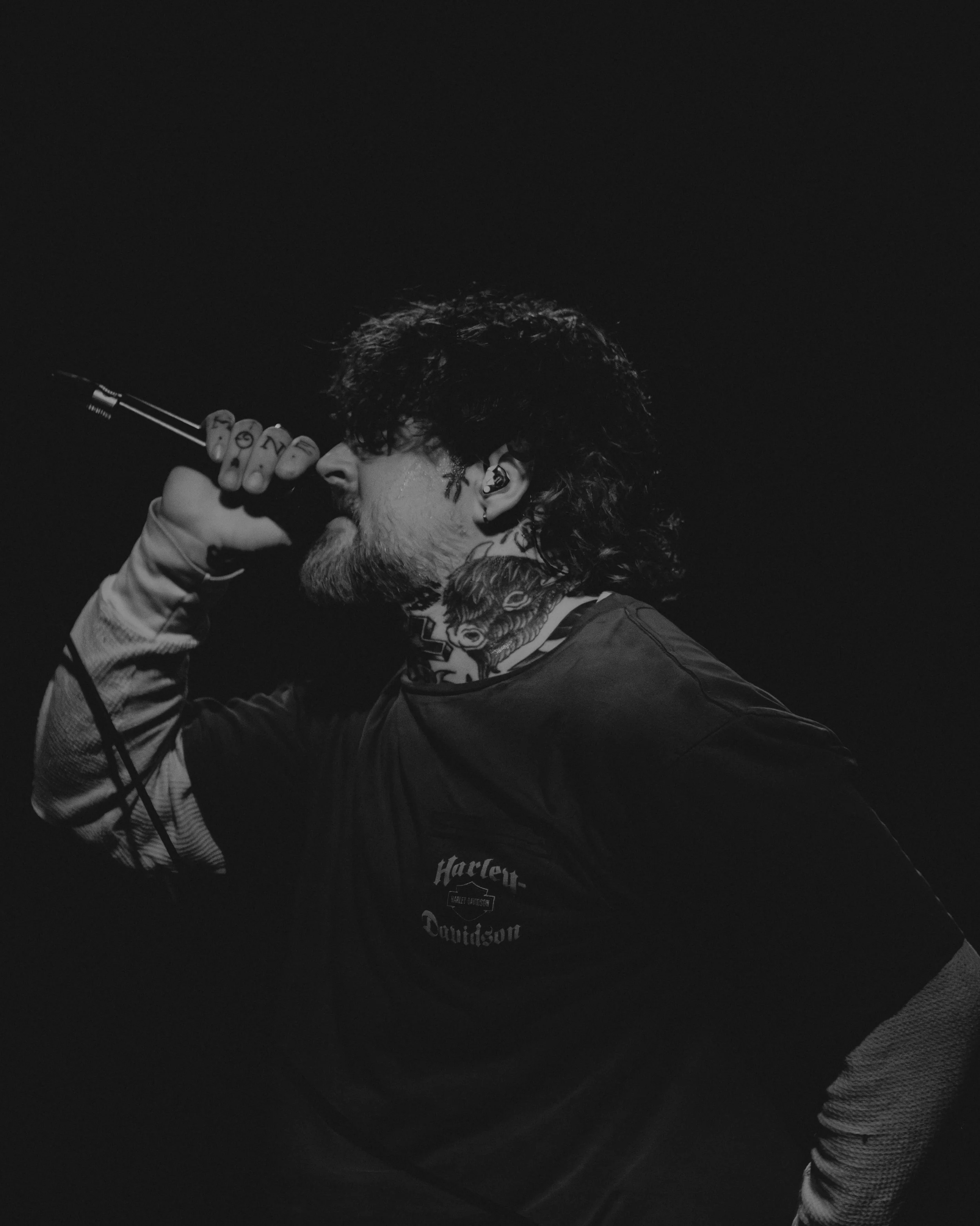 A person with tattoos on their neck and face, wearing a Harley-Davidson shirt, is holding a guitar close to their face, with their eyes closed and long curly hair falling to the side.