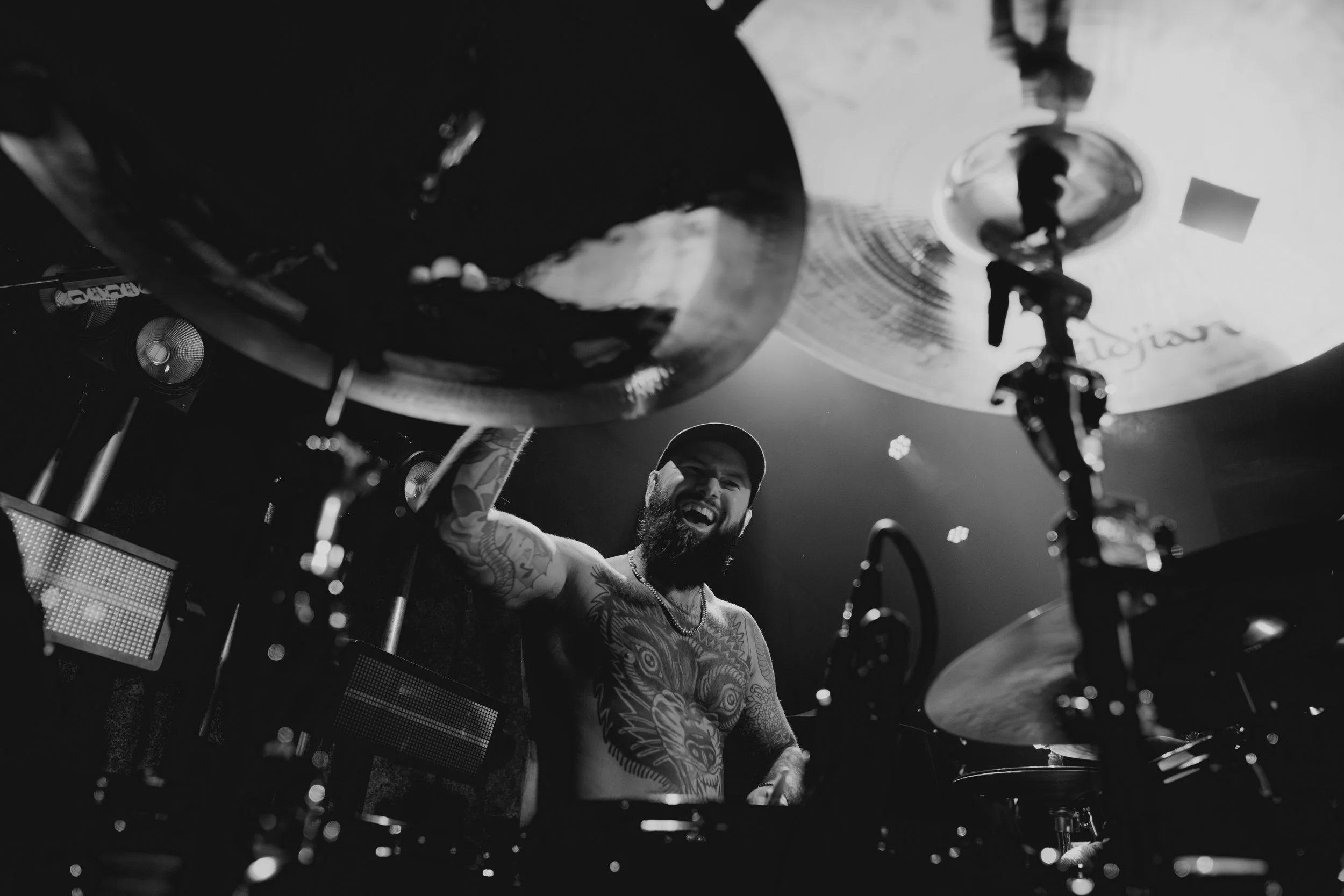 A man with tattoos playing the drums on stage, smiling, holding drumsticks, with a drum set and stage lights in the background.