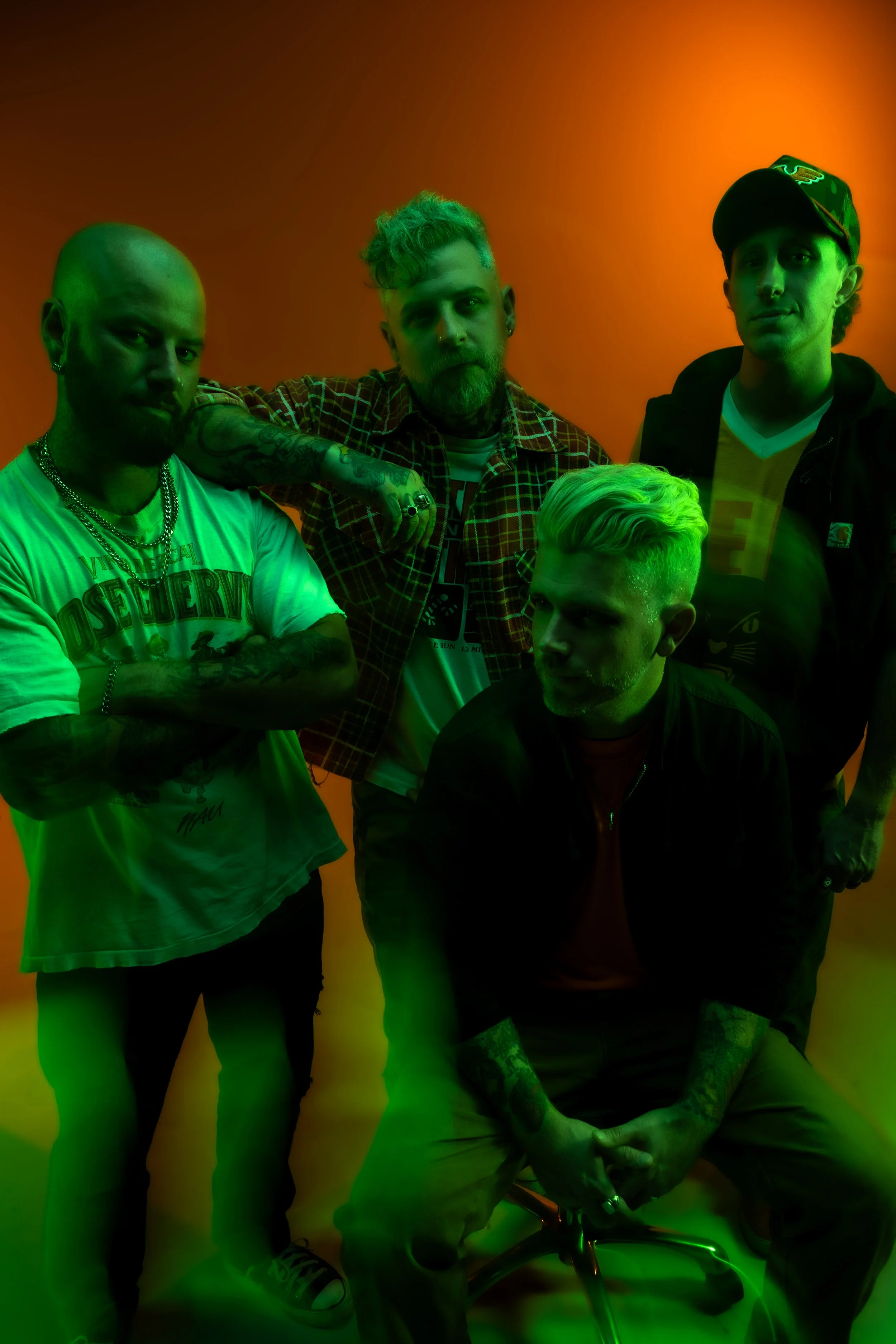 Four young men with tattoos, jewelry, and casual clothing, standing in front of a vibrant orange and green gradient background.