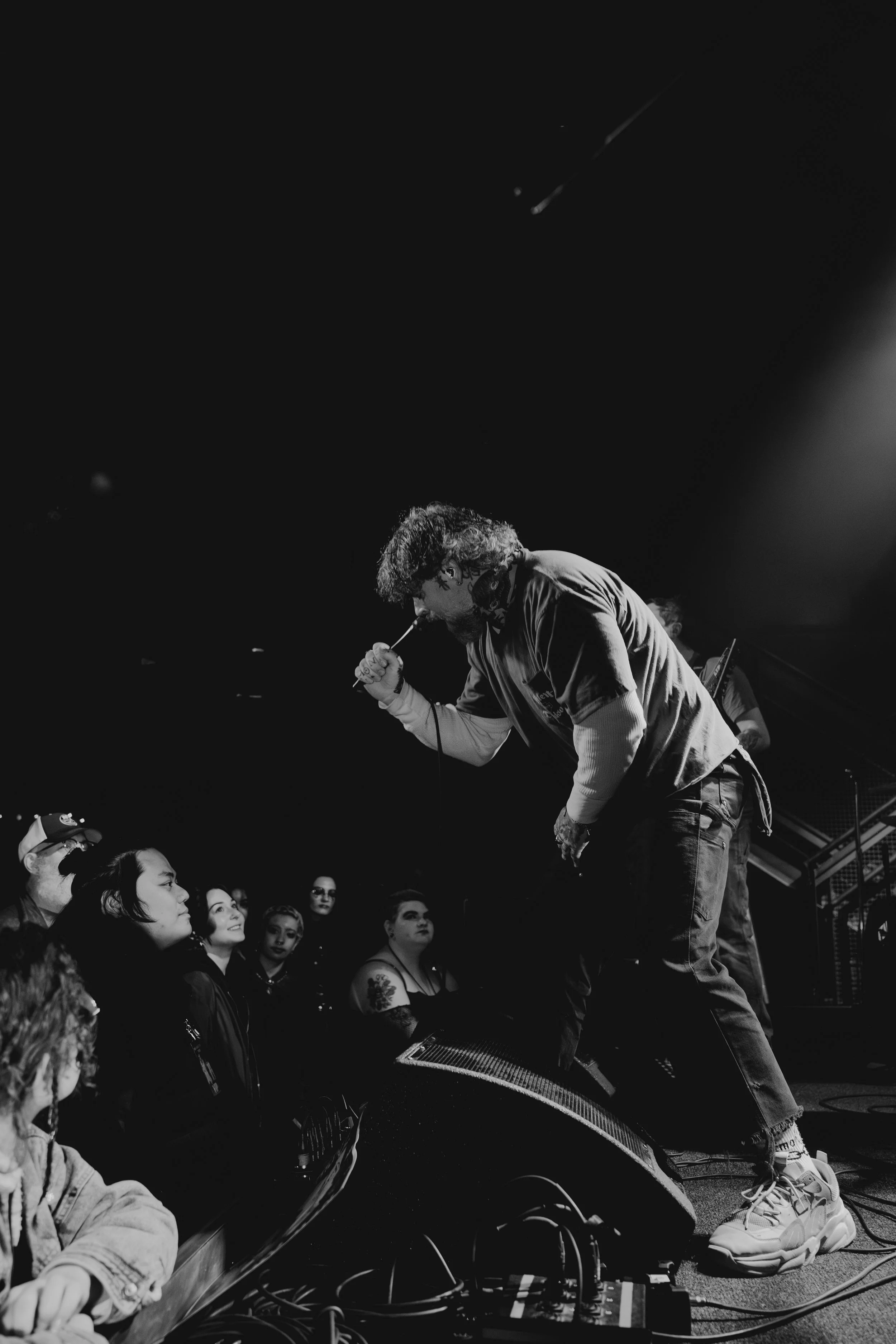 Male performer with curly hair and beard, wearing a casual T-shirt and baggy pants, leaning forward and singing into a microphone on stage in front of a seated audience at a concert.