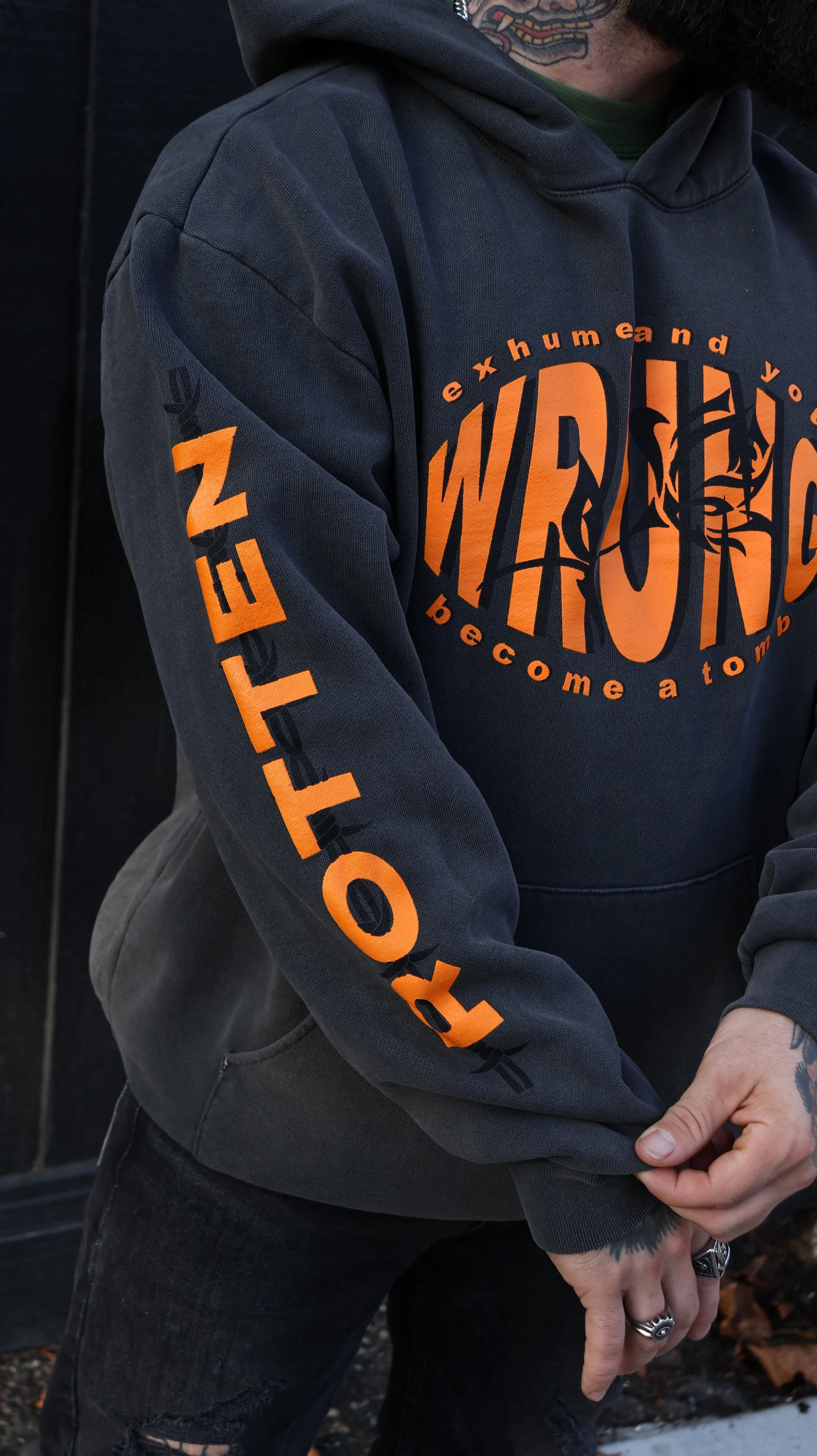A person wearing a black hoodie with orange text and graphics, with visible tattoos and rings, standing outdoors.