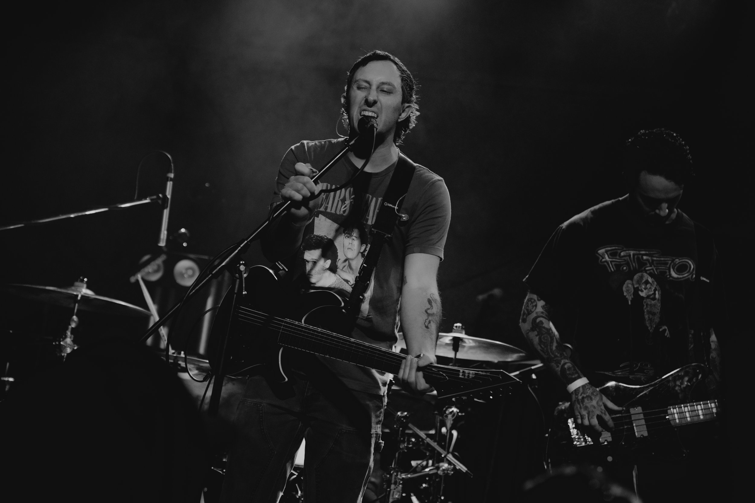 A black and white photo of a man singing into a microphone while playing guitar, with another musician playing bass guitar beside him and drums in the background.