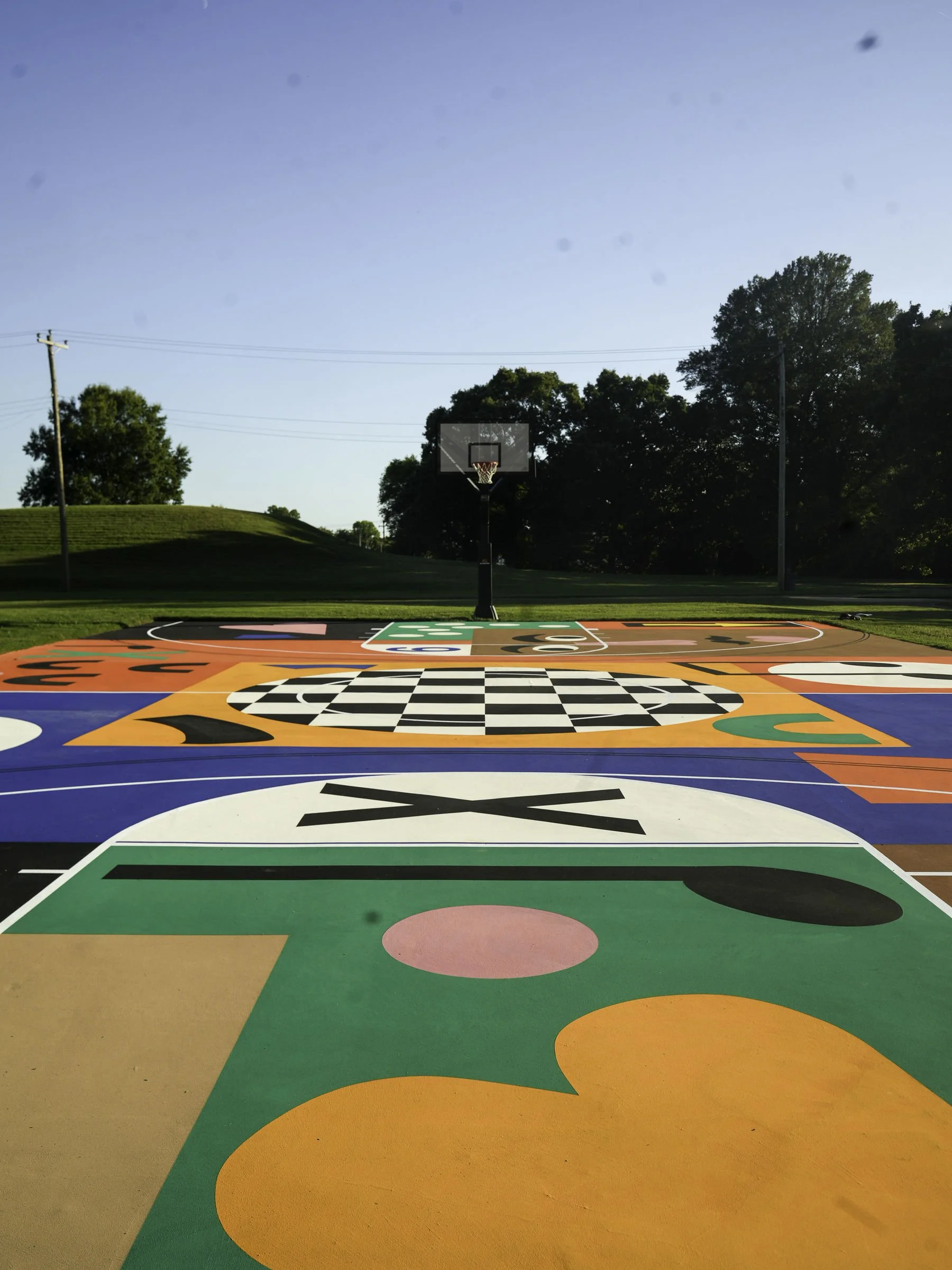 Colorful outdoor basketball court with various patterns and designs painted on the surface, set in a park with trees and clear blue sky in the background.