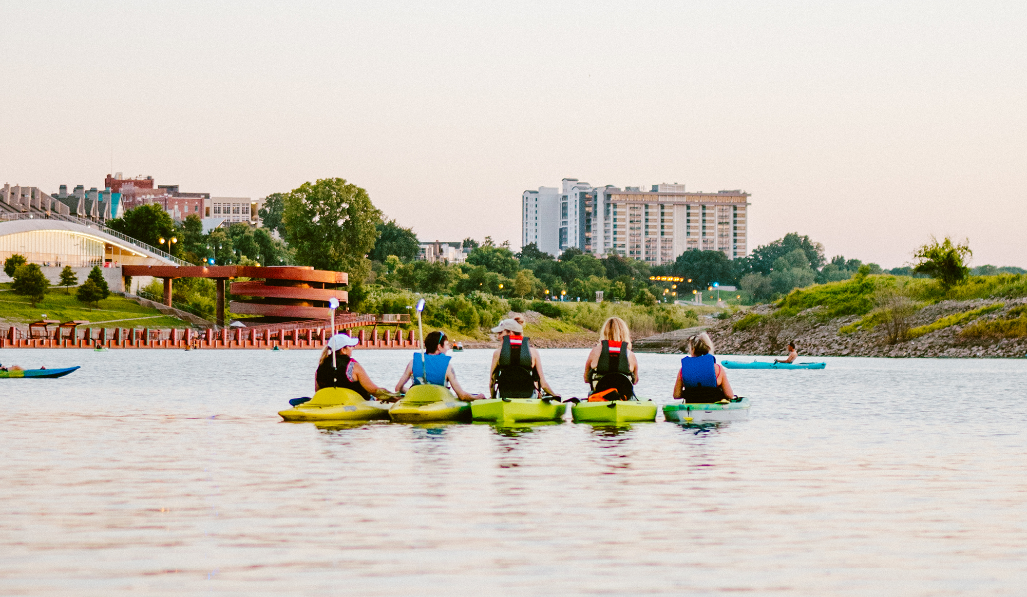 Group of people kayaking on a river with city buildings and trees in the background during sunset.