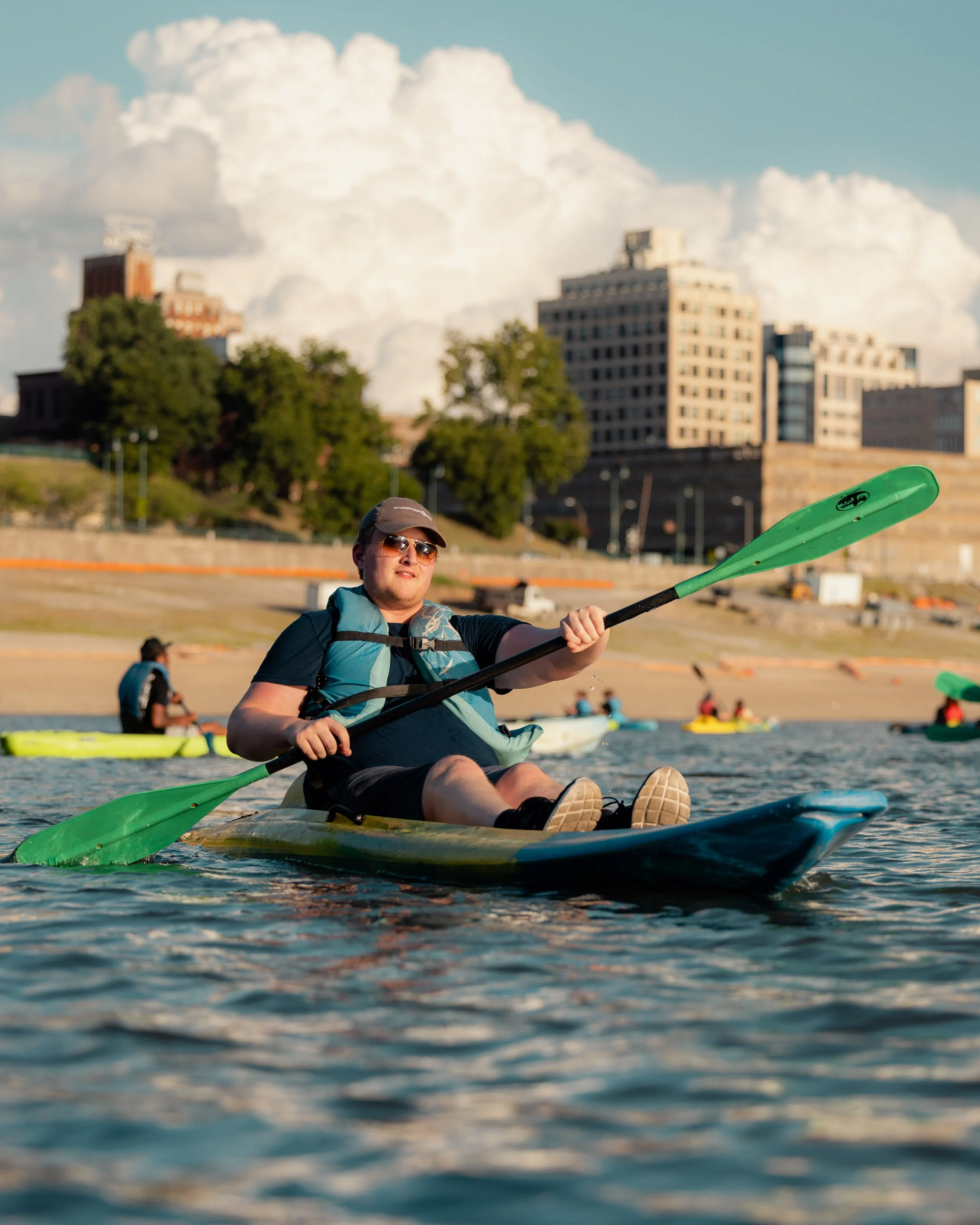 Person kayaking on a river with city buildings and cloudy sky in the background.