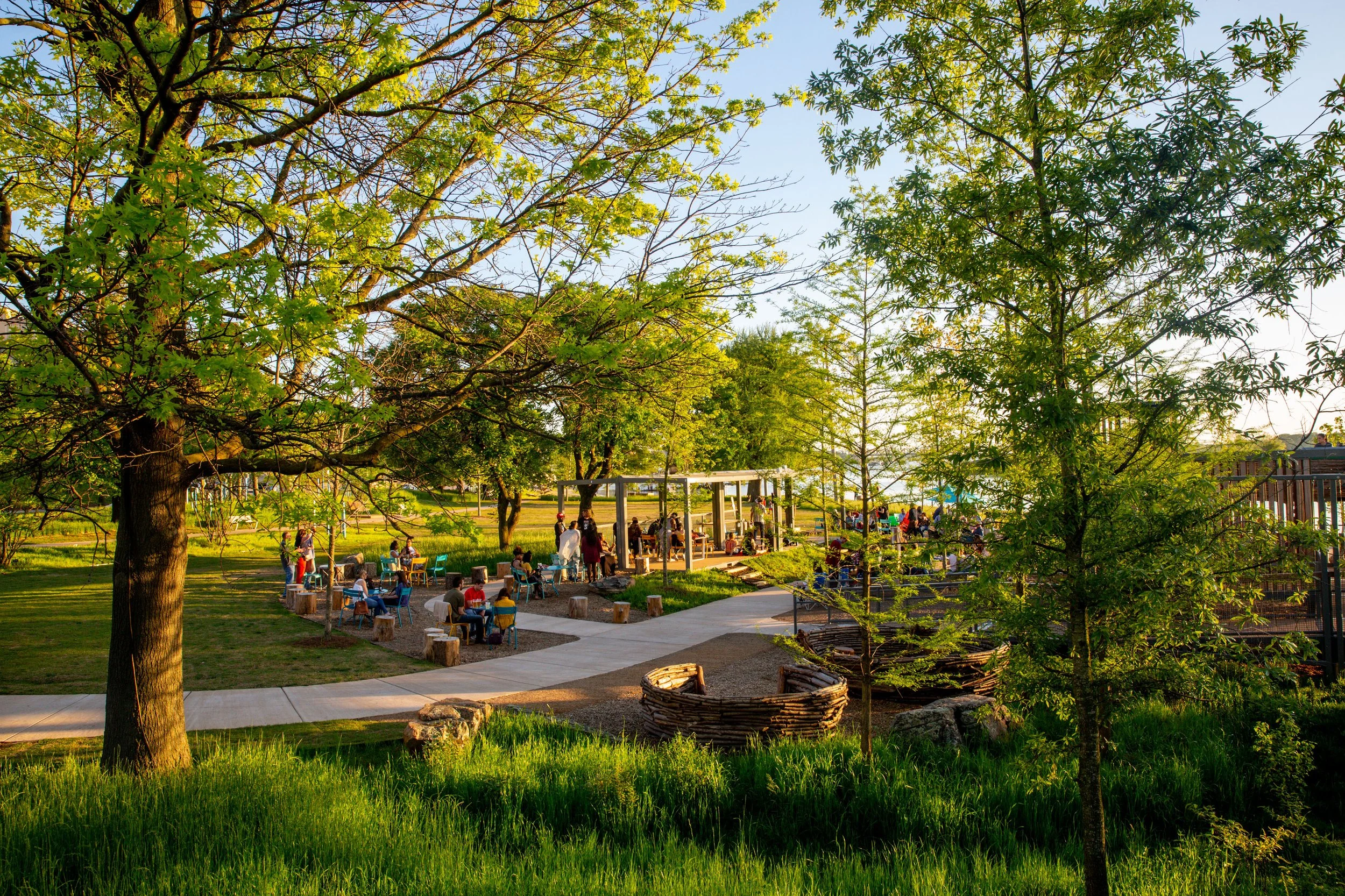 A park with tall trees and lush green grass. People are gathered around tables and under a shelter, enjoying the sunny day. A pathway runs through the park, and a woven basket is in the foreground.
