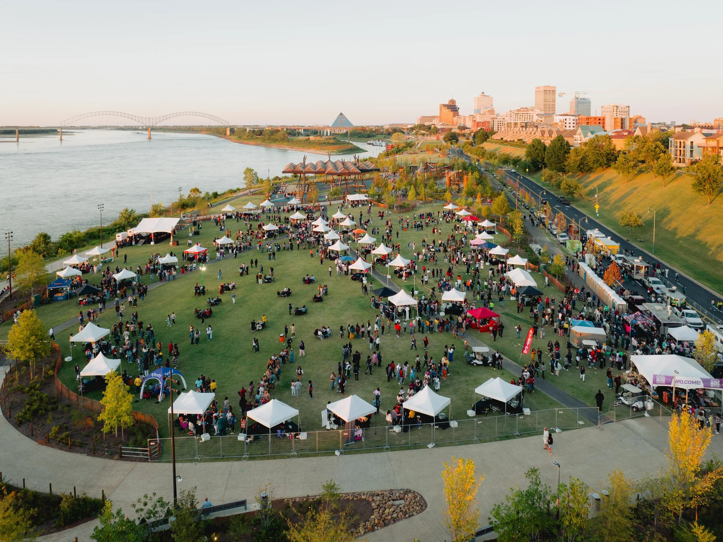 An aerial view of a cityscape with a river on the left, a park filled with tents and people in the foreground, and a downtown skyline with tall buildings in the background during sunset.