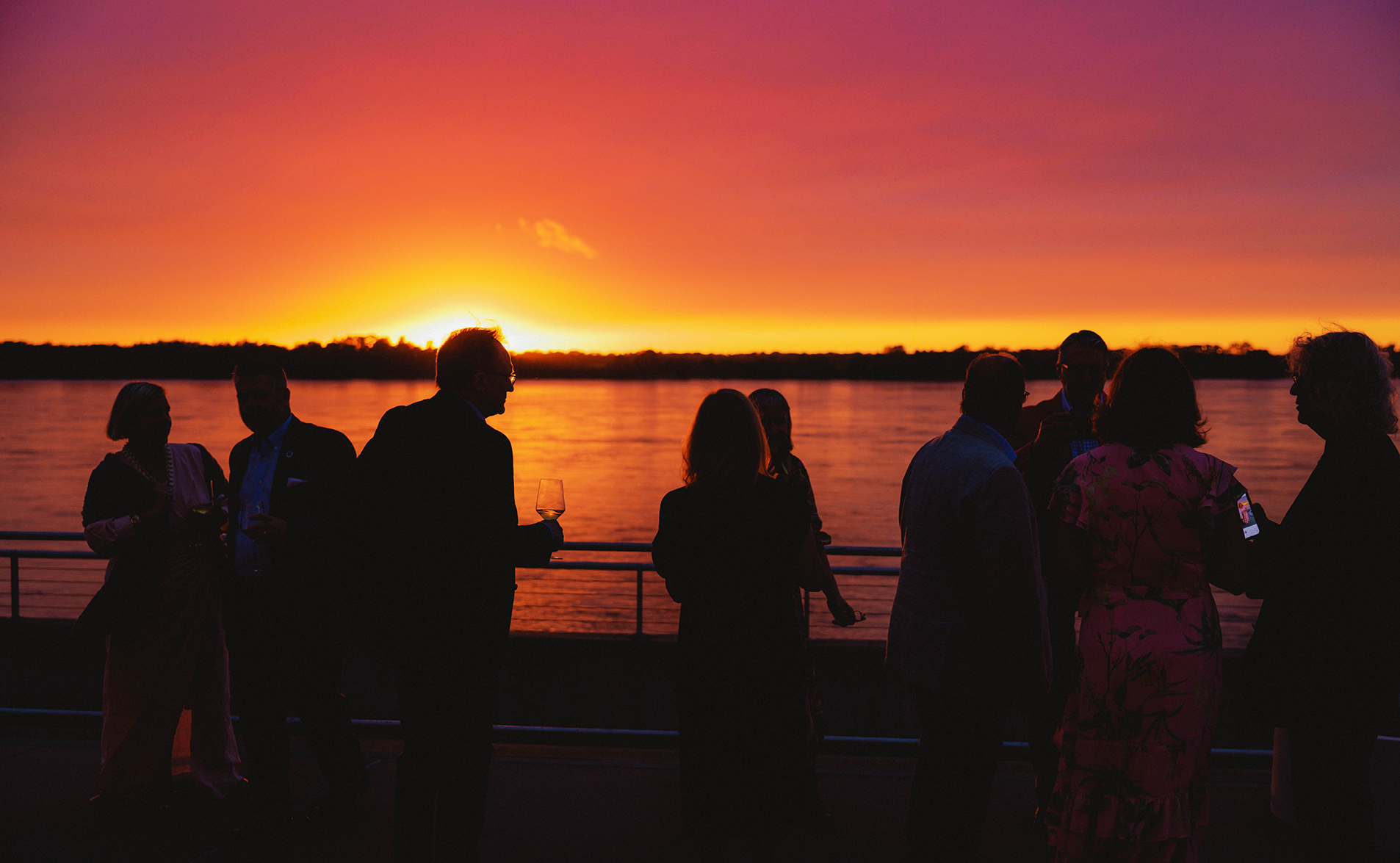 Silhouettes of people socializing on a boat during sunset, with orange and purple sky and water visible in the background.