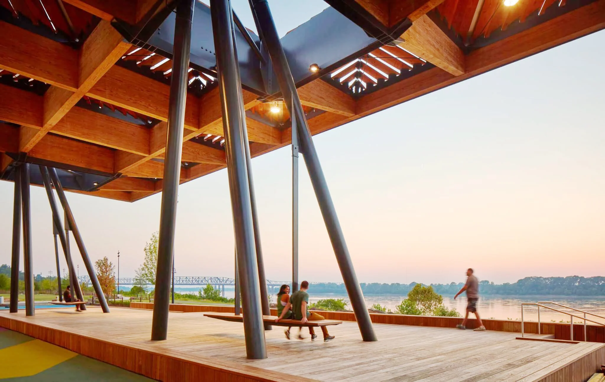 People sitting on a wooden deck under a modern wooden and steel pavilion near a river, with a bridge visible in the background during sunset.