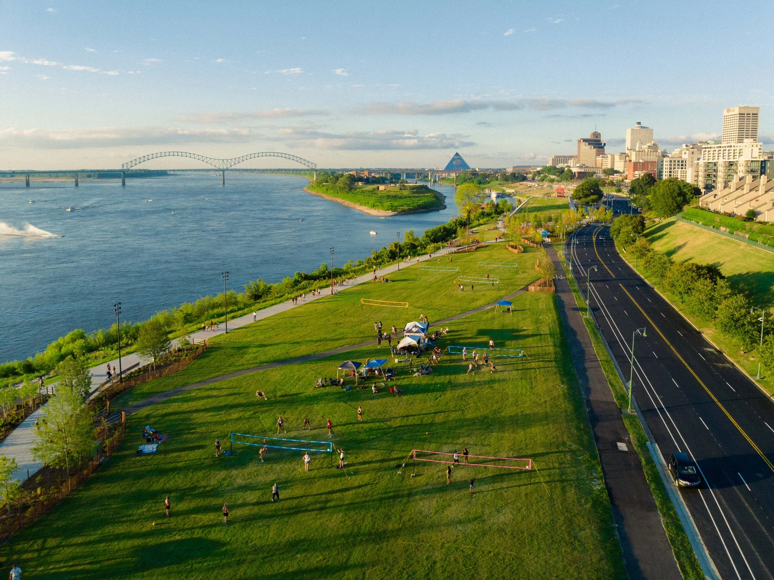 A scenic cityscape with a river, a bridge, and a skyline of tall buildings. A park with people playing volleyball and walking along paths is in the foreground, with a stadium and other green spaces.