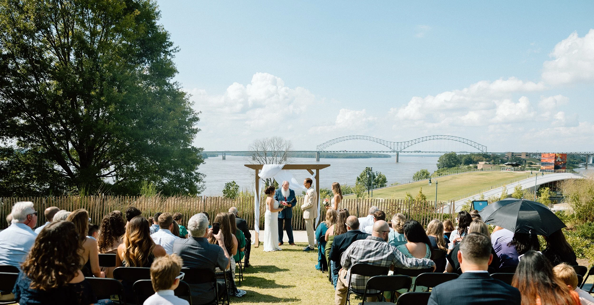 Outdoor wedding ceremony taking place near a river with a bridge in the background, guests seated on chairs, and the couple exchanging vows under a simple arch.