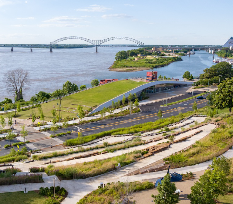 View of a river with a bridge in the background, a landscaped park with walking paths, green lawns, trees, and a modern building underneath a curved roof structure.