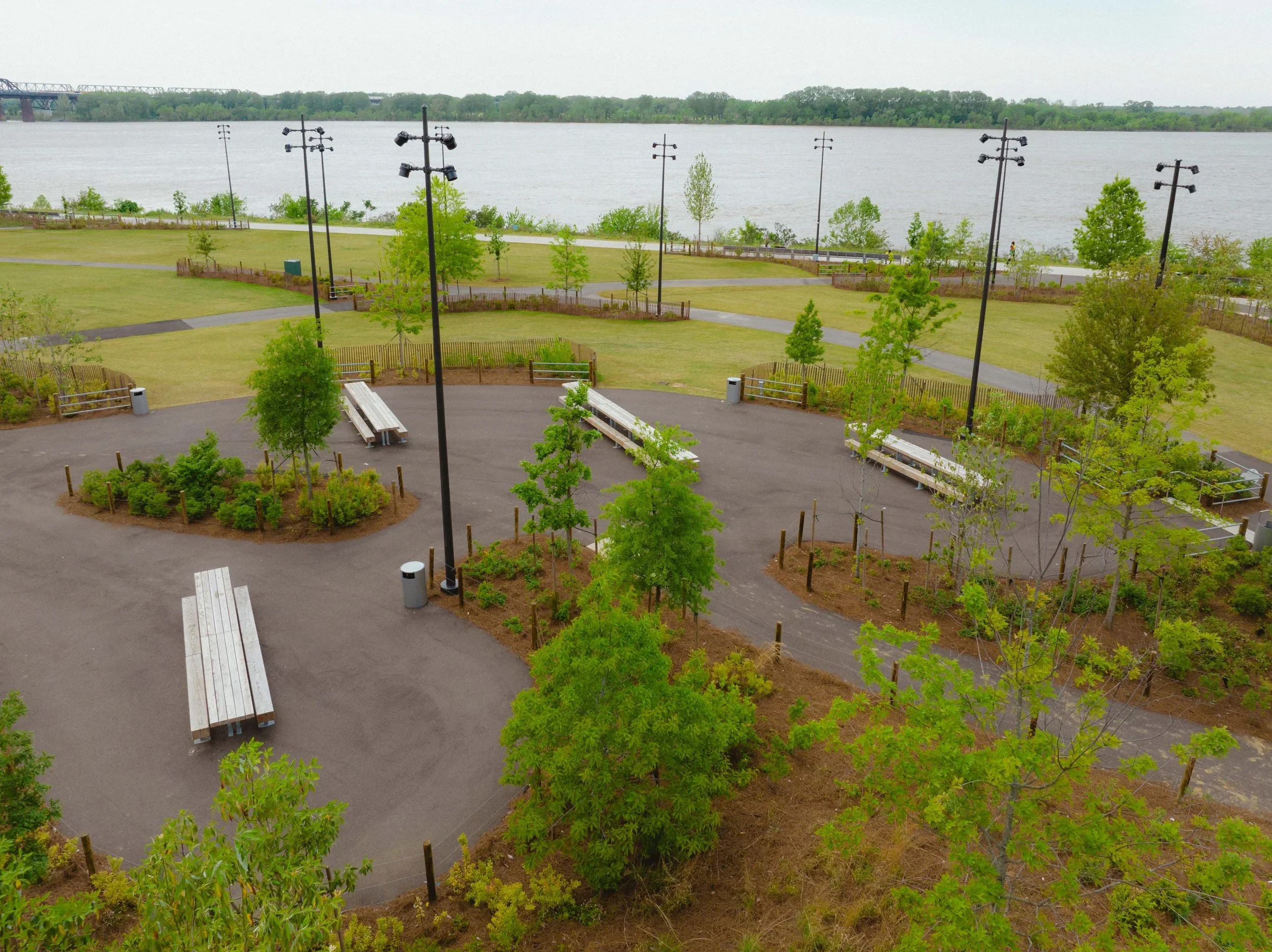 A park with walking paths, benches, young trees, and tall street lamps near a large body of water with a bridge in the background.