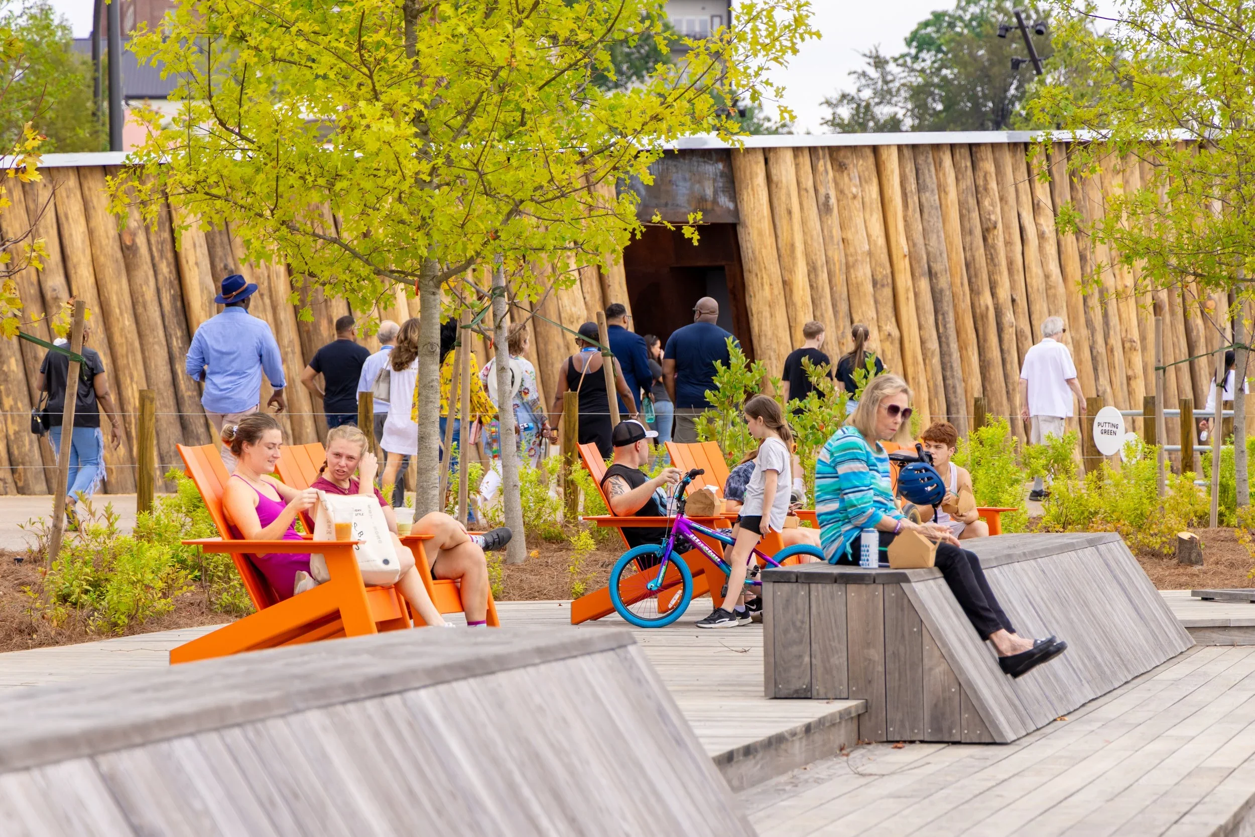 People sitting and walking in a park with orange benches, trees, and a wooden wall in the background.
