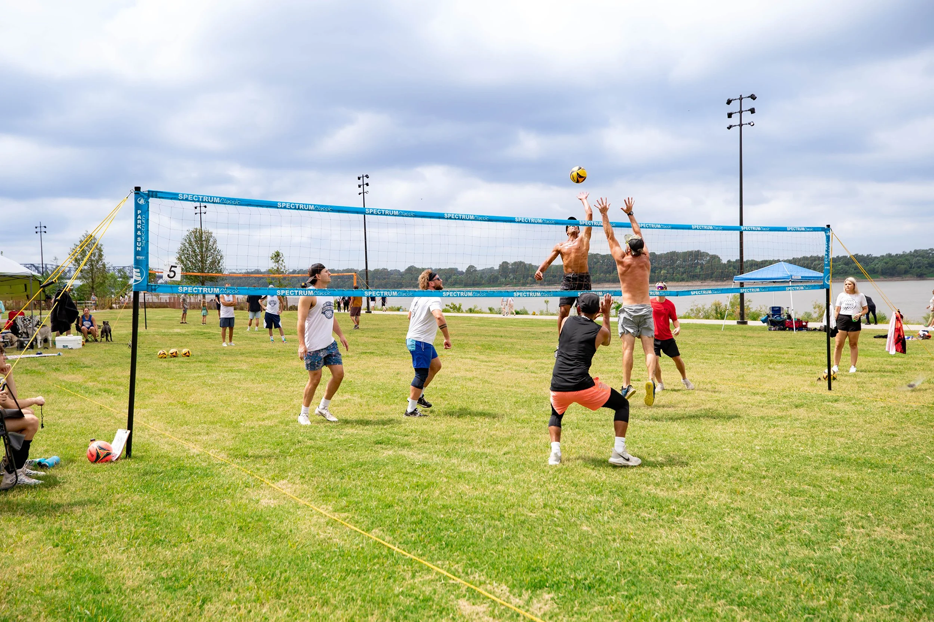 People playing volleyball outdoors near a body of water on a cloudy day, with some spectators and a tent nearby.