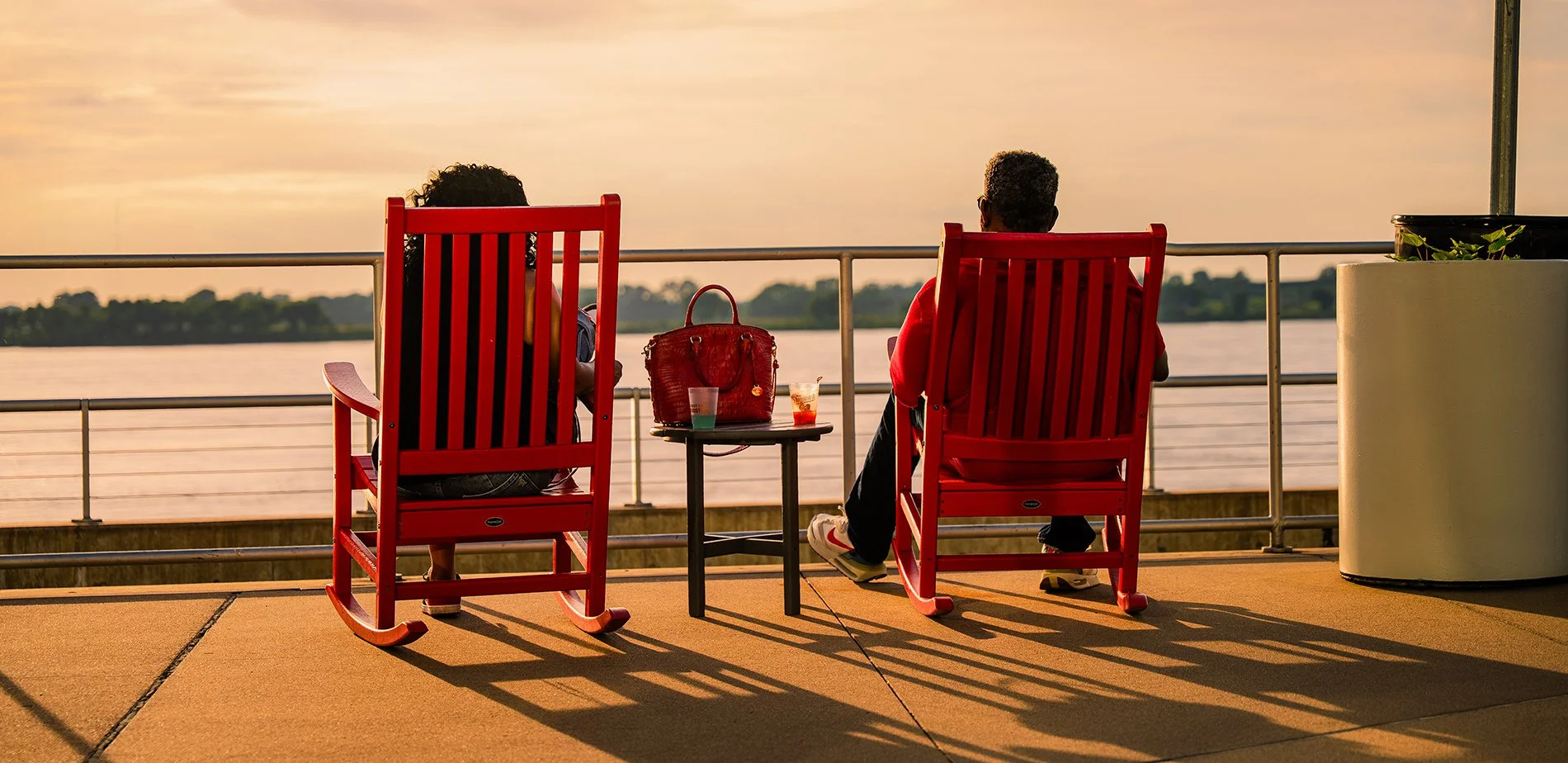 Two people sitting on red rocking chairs on a waterfront promenade at sunset, with a small table between them holding a red purse and drinks, overlooking a river and distant land.