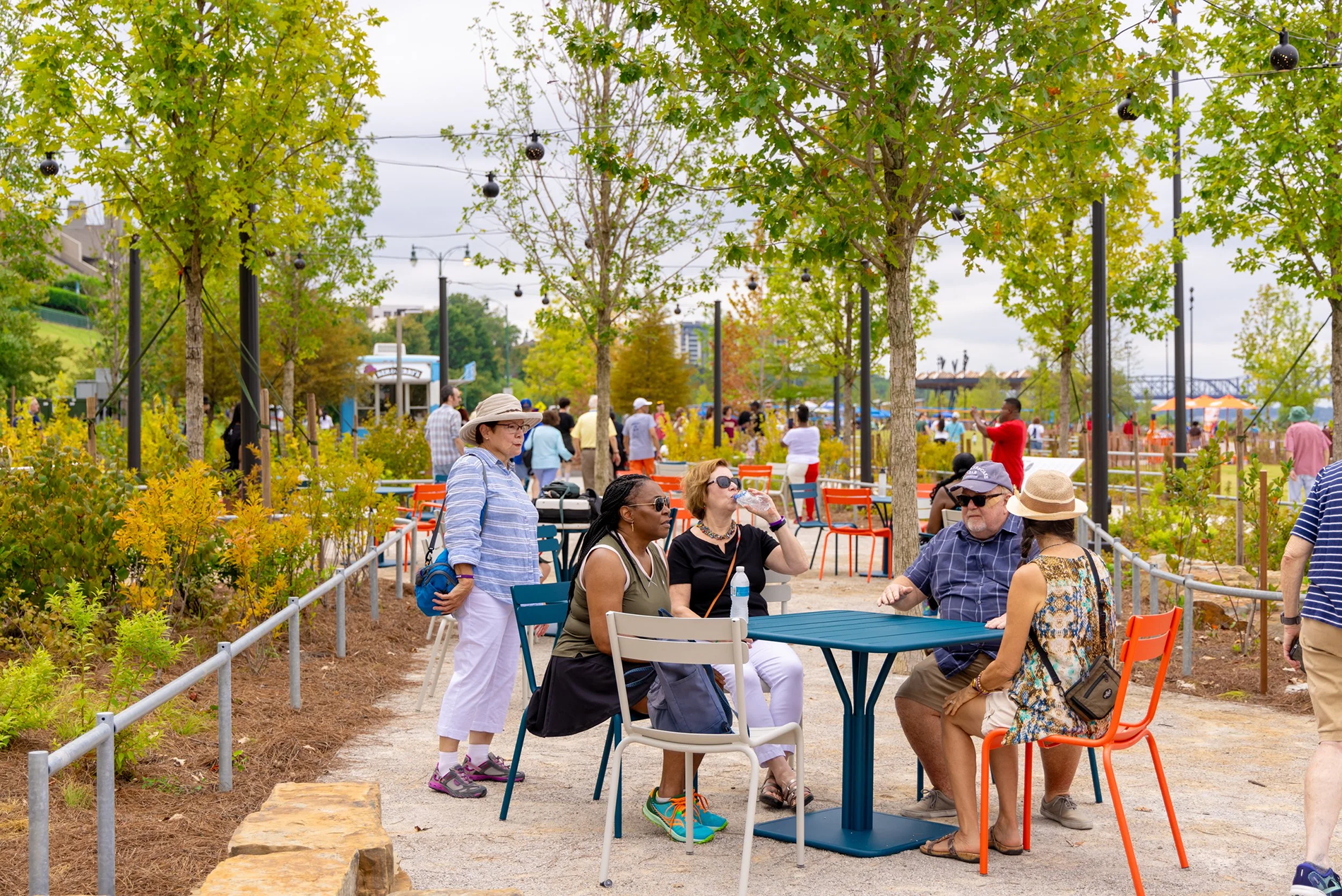 People sitting at outdoor tables and standing in a park-like setting with green trees, string lights, and a cloudy sky.