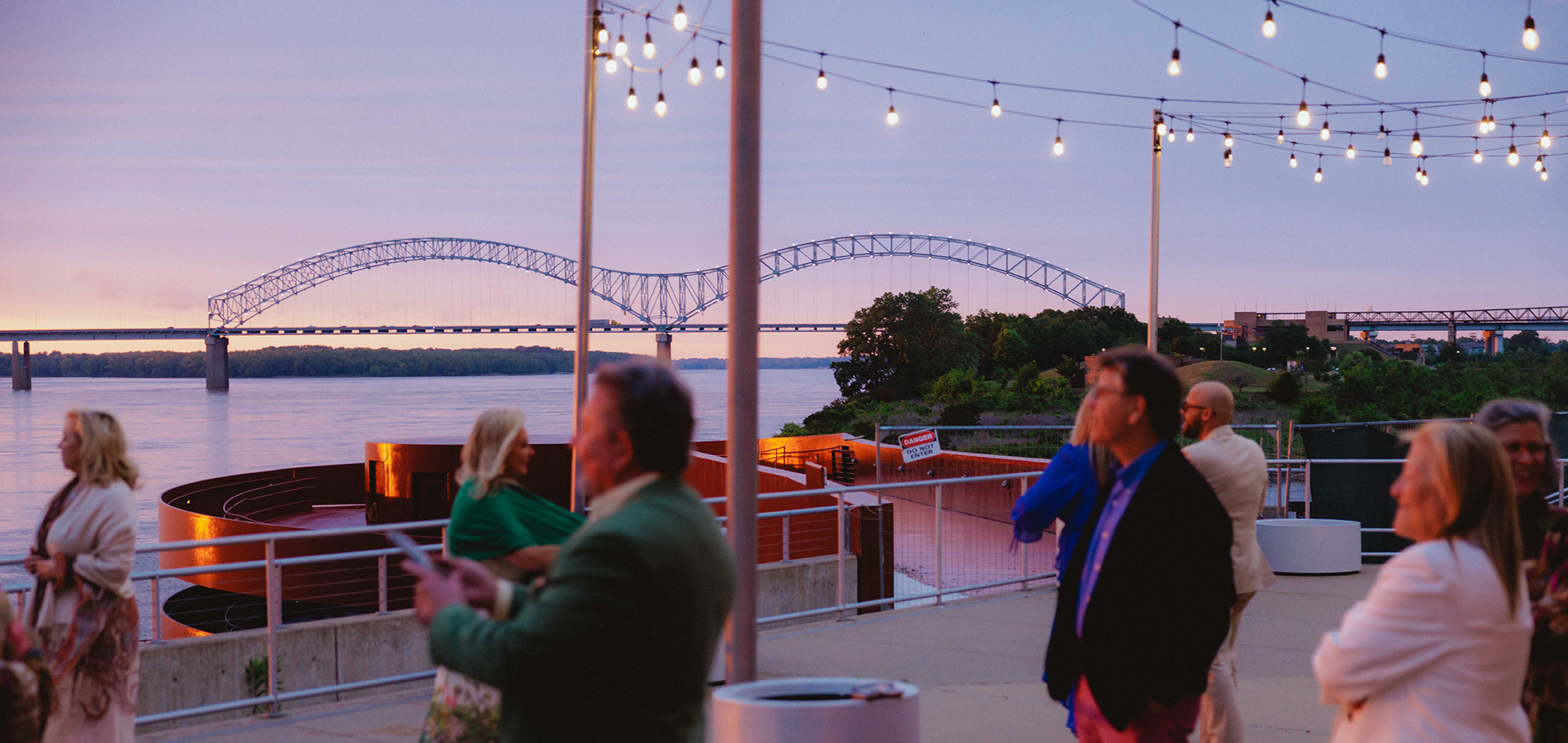 People socializing on a deck during sunset with a bridge in the background and string lights overhead.