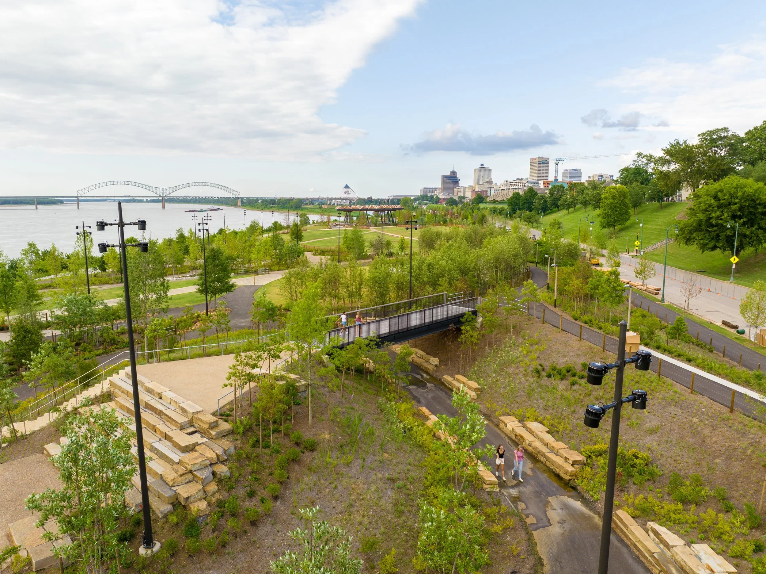 View of a park overlooking a river with a city skyline in the background, including a bridge and various buildings, with green trees, walking paths, and a few people strolling.