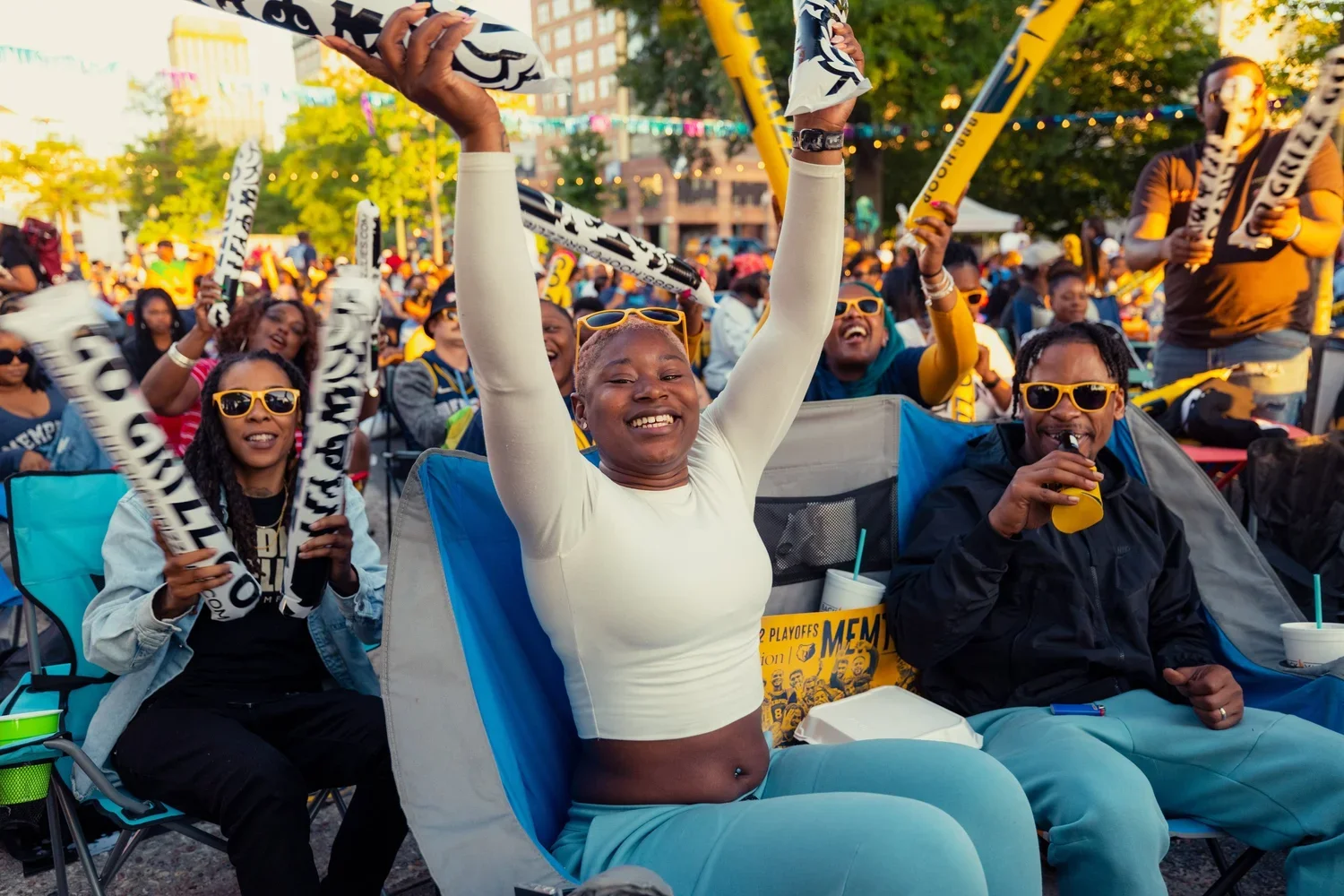 Crowd of excited sports fans cheering and enjoying the game outdoors, some wearing yellow sunglasses, with flags and banners, during a sunny day.