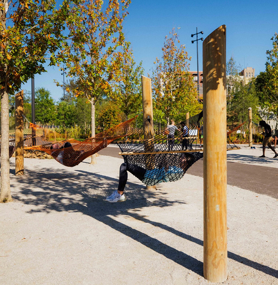 People relaxing in hammocks set between wooden posts in a park with trees and a blue sky.