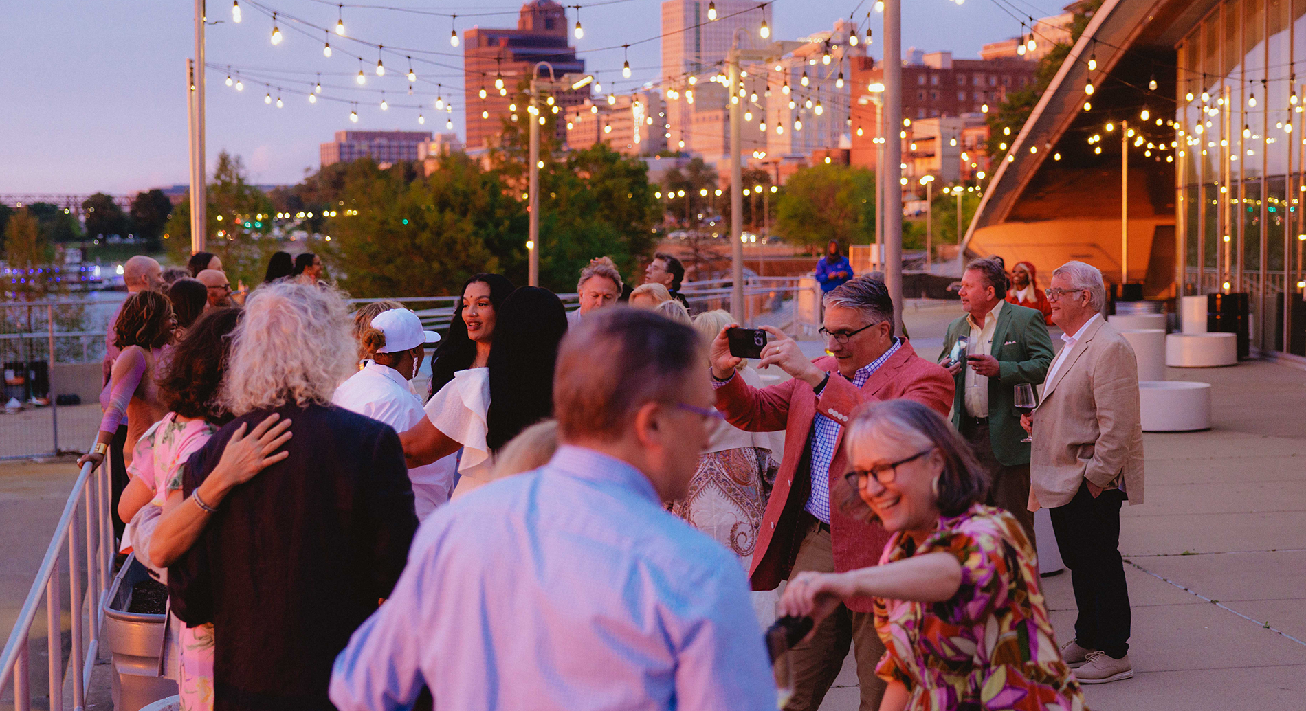 People socializing outdoors on a deck during evening, with string lights and cityscape in the background.