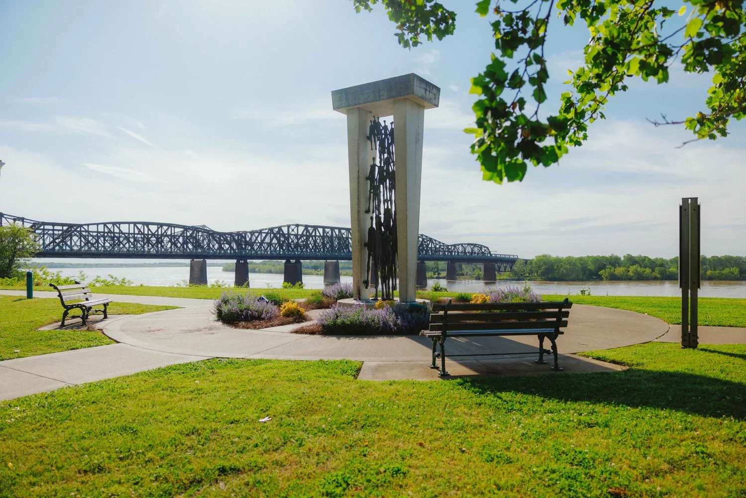 A park by a river features a sculpture of hanging human figures under a concrete arch, with benches, a pathway, and lush greenery. A bridge spans the river in the background.