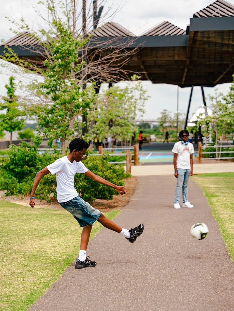A boy in a white t-shirt and denim shorts kicking a soccer ball on a park pathway, with a girl nearby and other people in the background on a sunny day.
