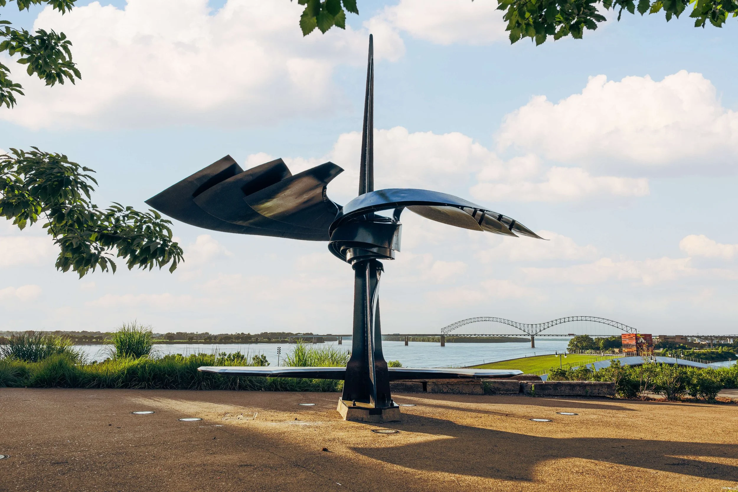 Outdoor sculpture situated on a paved area near a river, with a bridge in the distance and trees with green leaves.