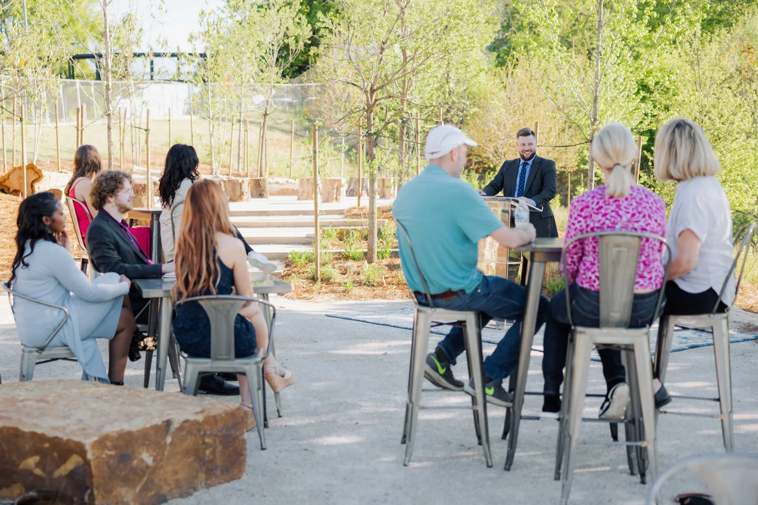A group of people attending an outdoor seminar or workshop in a lush, green park with trees, with a speaker smiling at a podium.