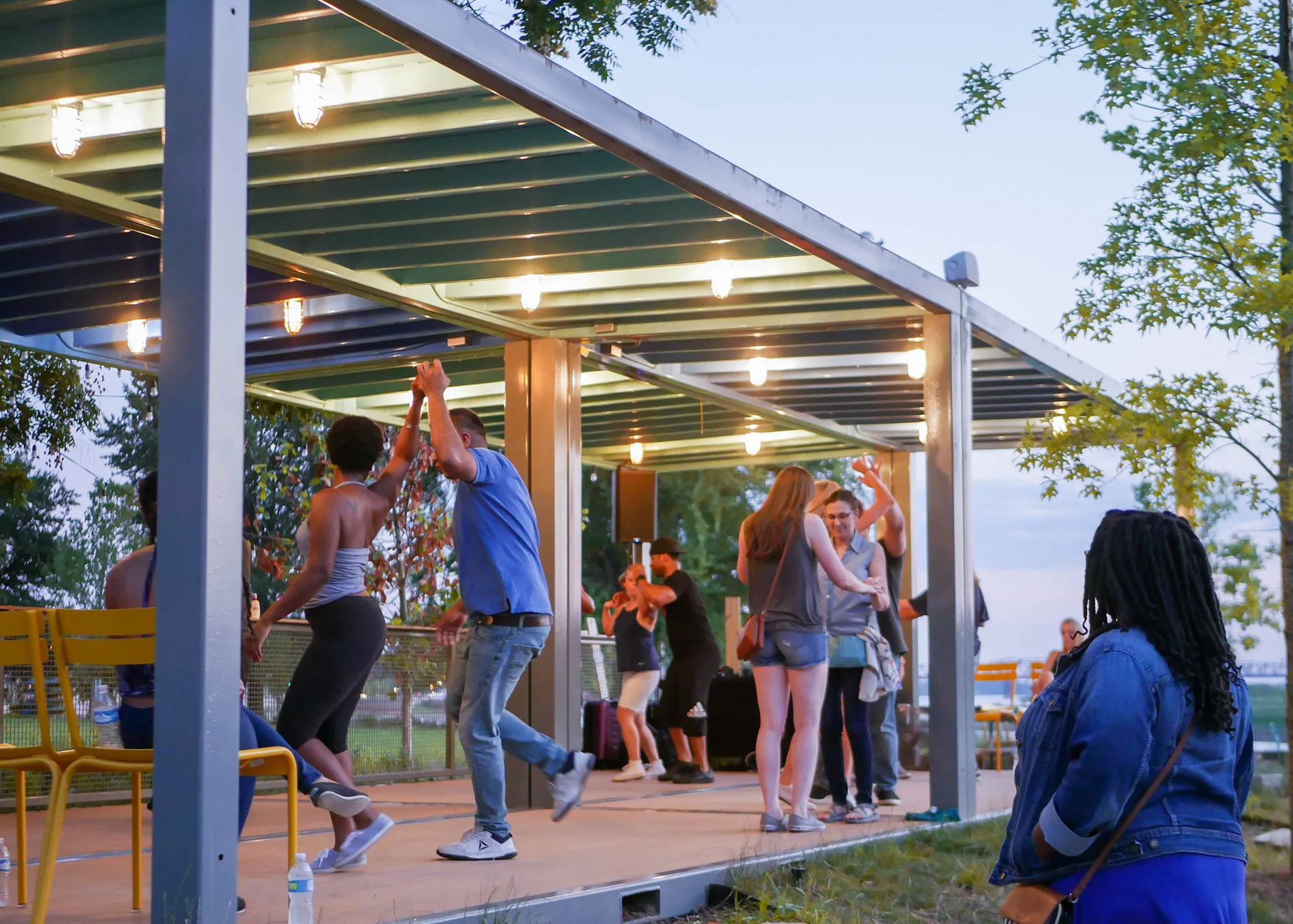 People dancing on an outdoor stage with string lights, surrounded by trees, during dusk.