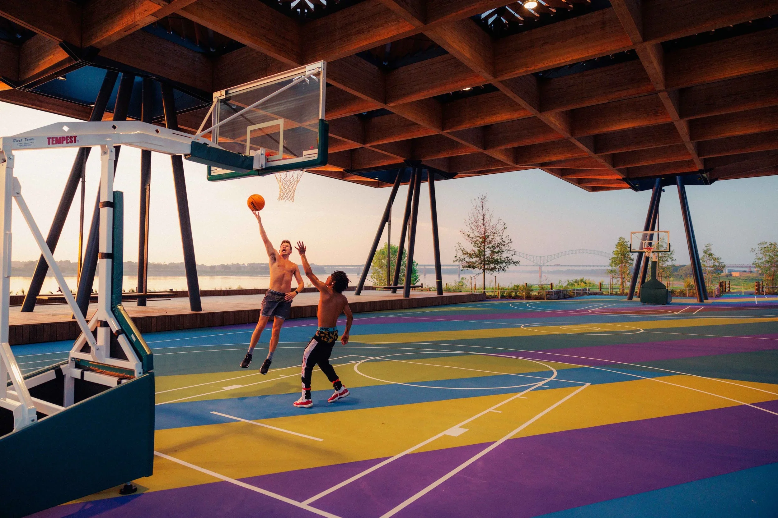 Two children playing basketball on an outdoor court under a large wooden roof structure, with a scenic river view and a bridge in the background during sunset.