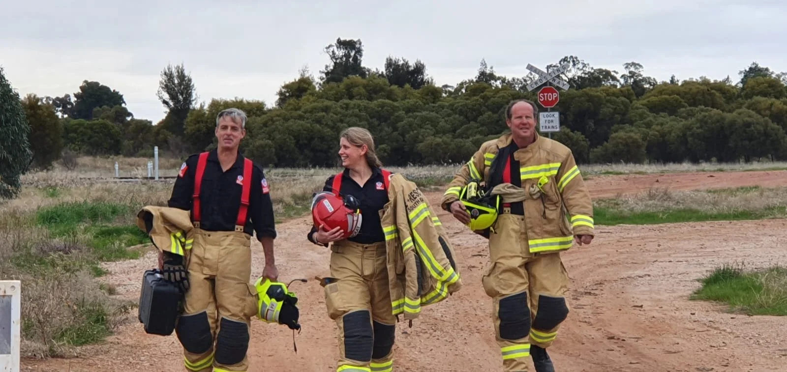 Three firefighters with Danielle McKay in the middle walking on a dirt road with trees in the background, carrying their helmets and gear, and smiling.