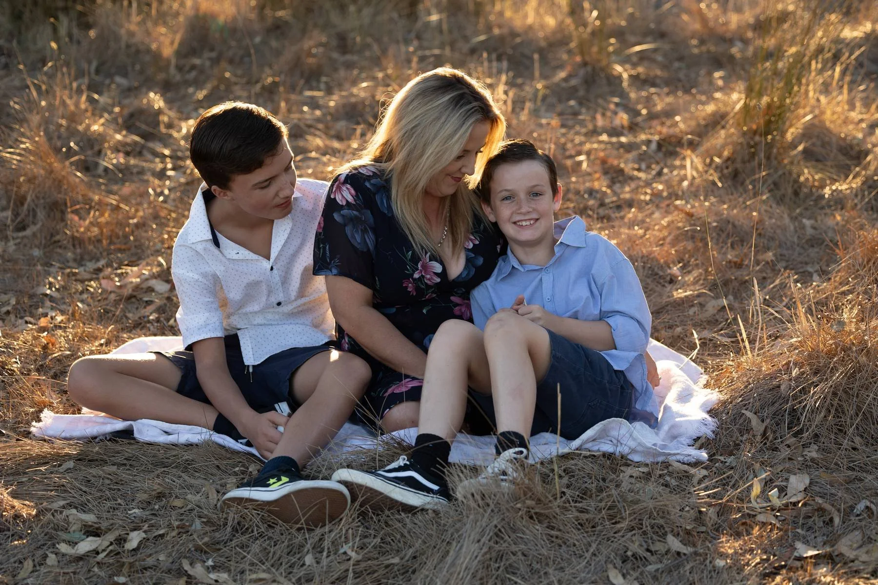 A photo taken by Danielle McKay Photography during a sunset family session of a mother and her two sons sitting on a blanket in a field of dry grass, smiling and enjoying each other's company during golden hour.