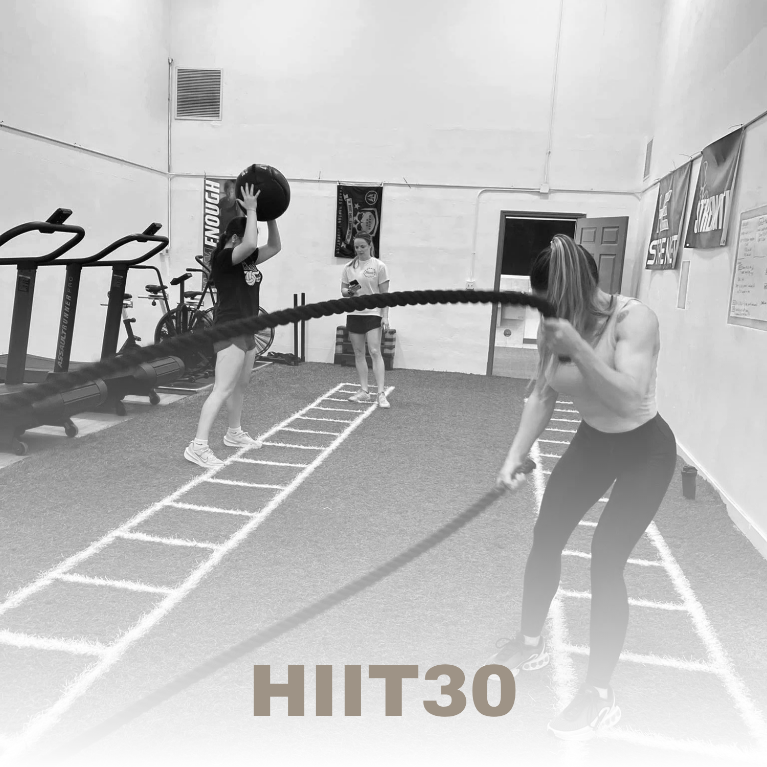 Two women exercising in a gym, one doing a battle rope workout and the other lifting a medicine ball, with a trainer standing in the background, in black and white.