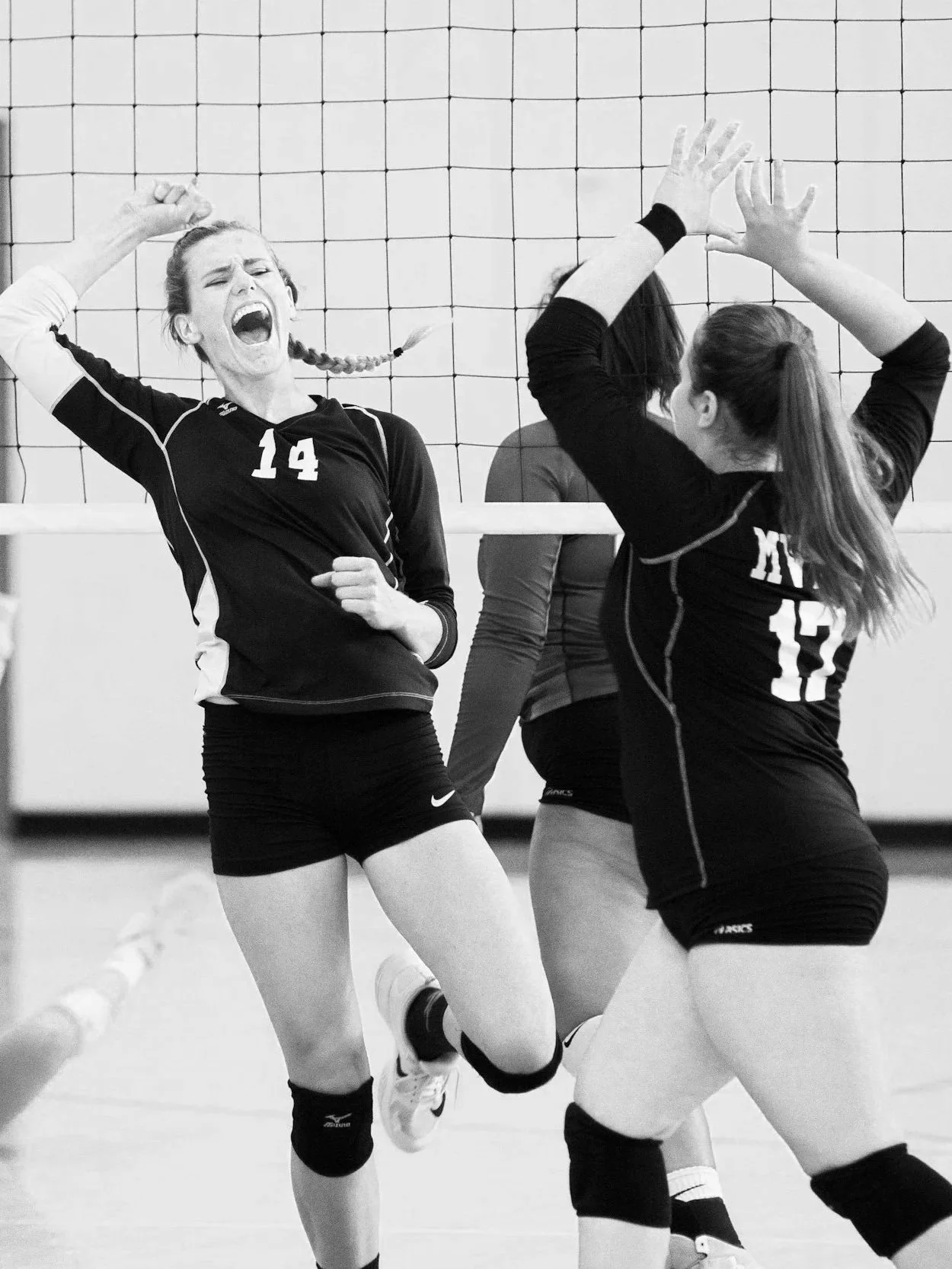 Three female volleyball players celebrating on the court, one with her mouth open and arms raised, two with arms up in congratulation, volleyball net in the background, black and white photo.